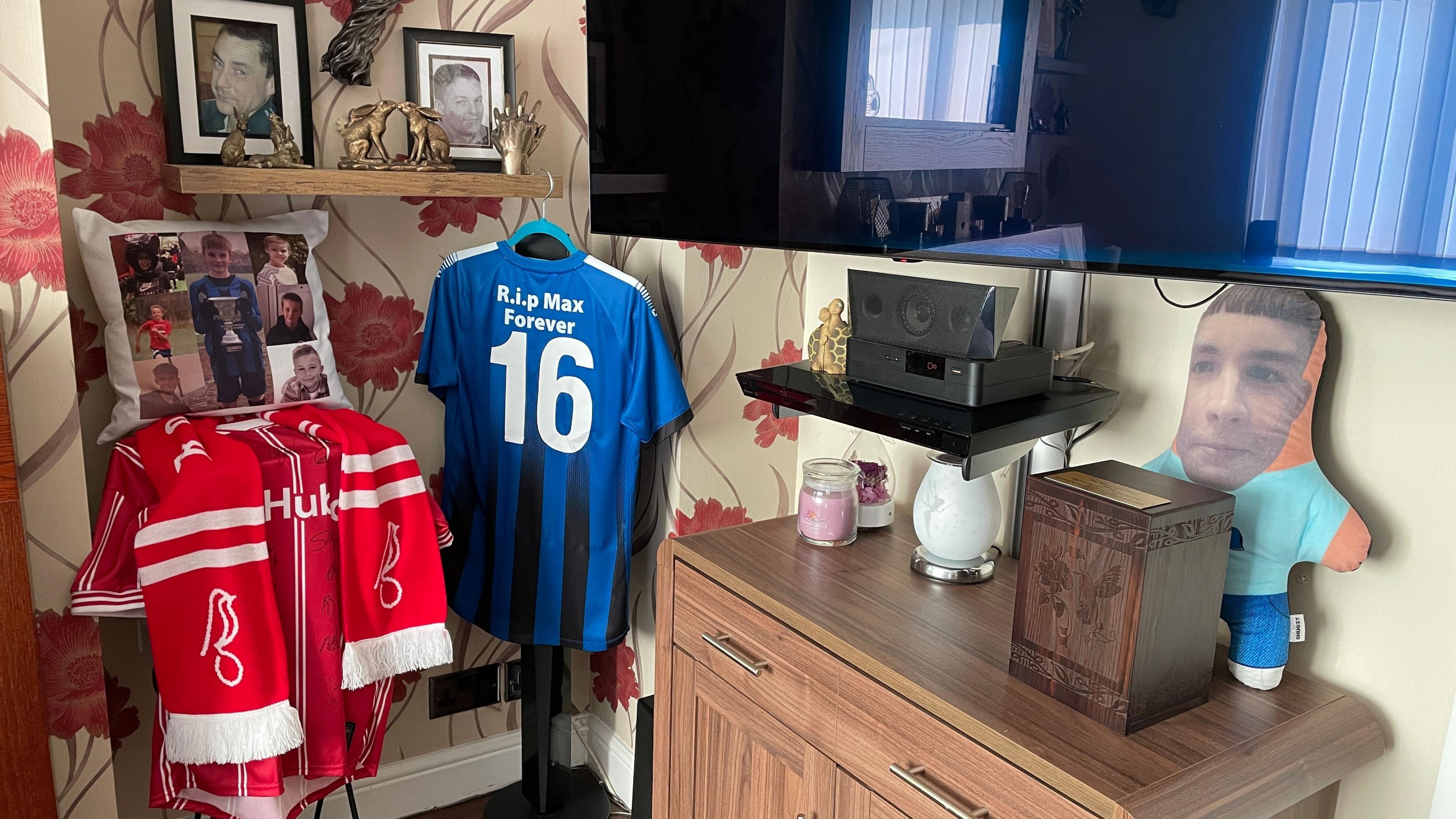 A cushion with pictures of teenager Max Dixon next to football shirts and a television. There is also a teddy with Max's face on it.