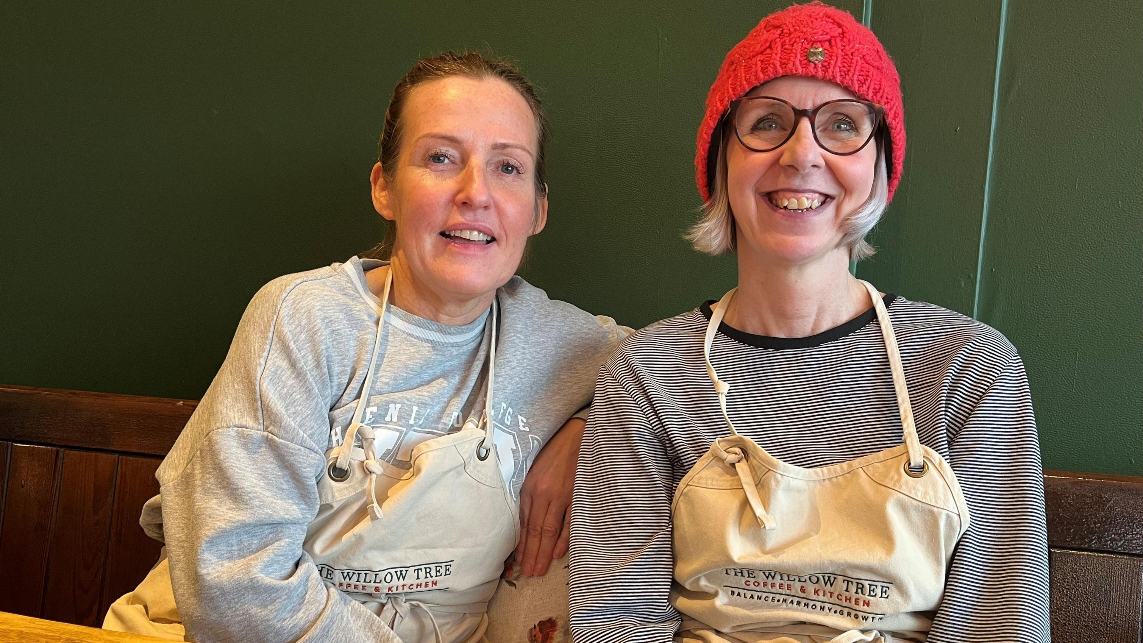 Katrina Brown and Jo Gordon sitting nest to each other and smiling at the camera. Katrina's blonde straight hair is tied at the back. She is wearing a light grey sweatshirt. 