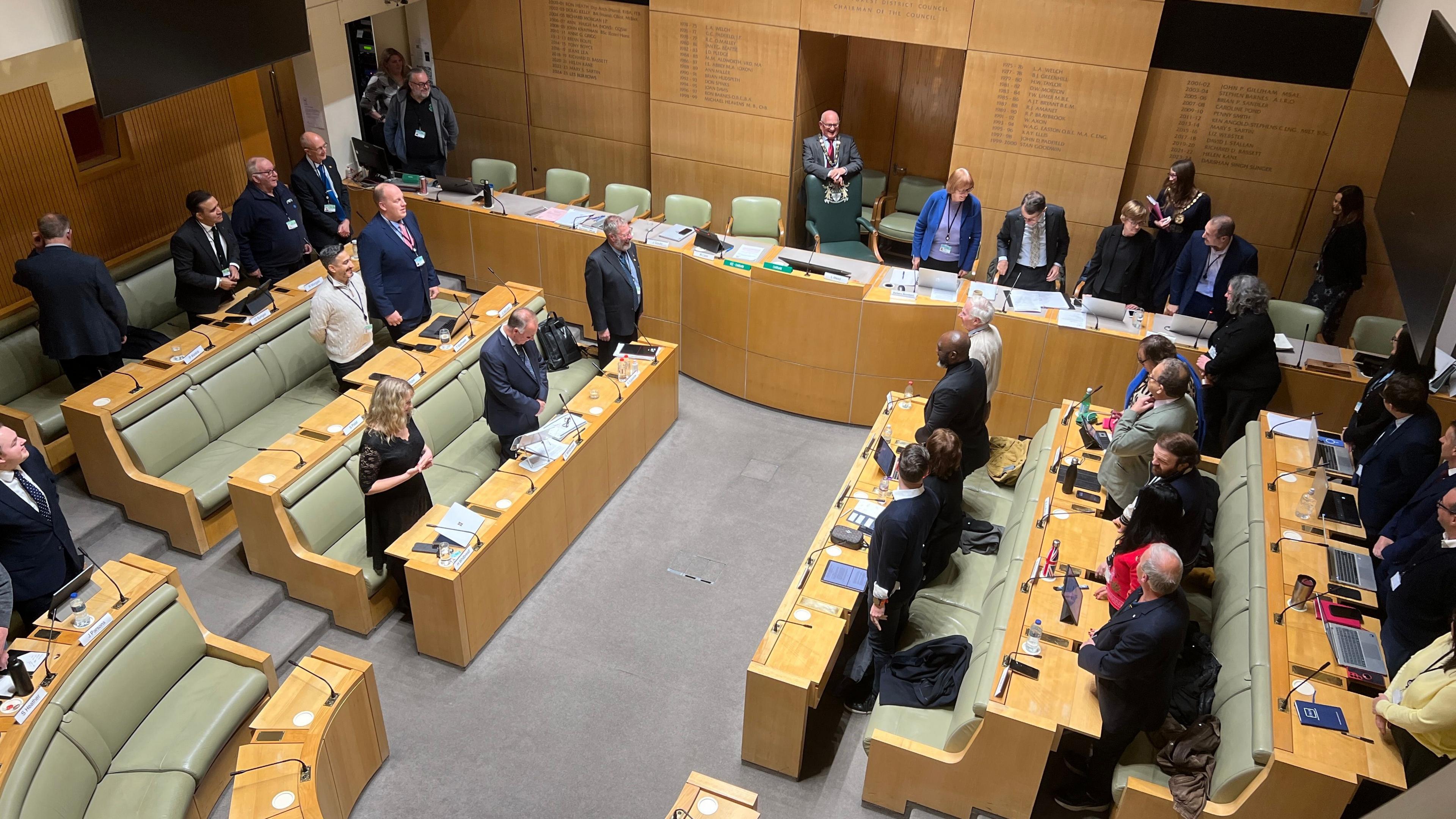 The council chamber, with councillors standing at the start of the meeting. There are many light green leather seats in front of desks with microphones.