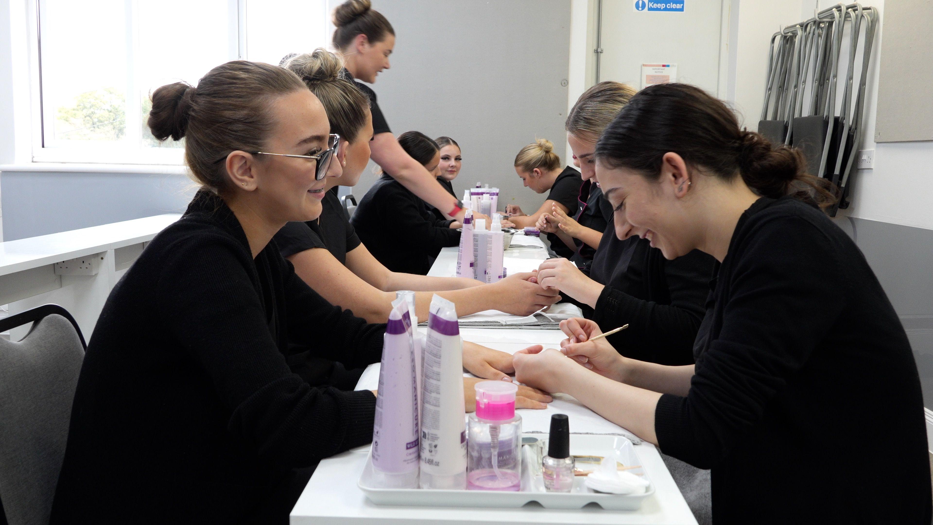 Female students sit either side of a long table in a beauty studio. They are either painting nails or having their nails painted. They all have their hair tied up and are wearing black.