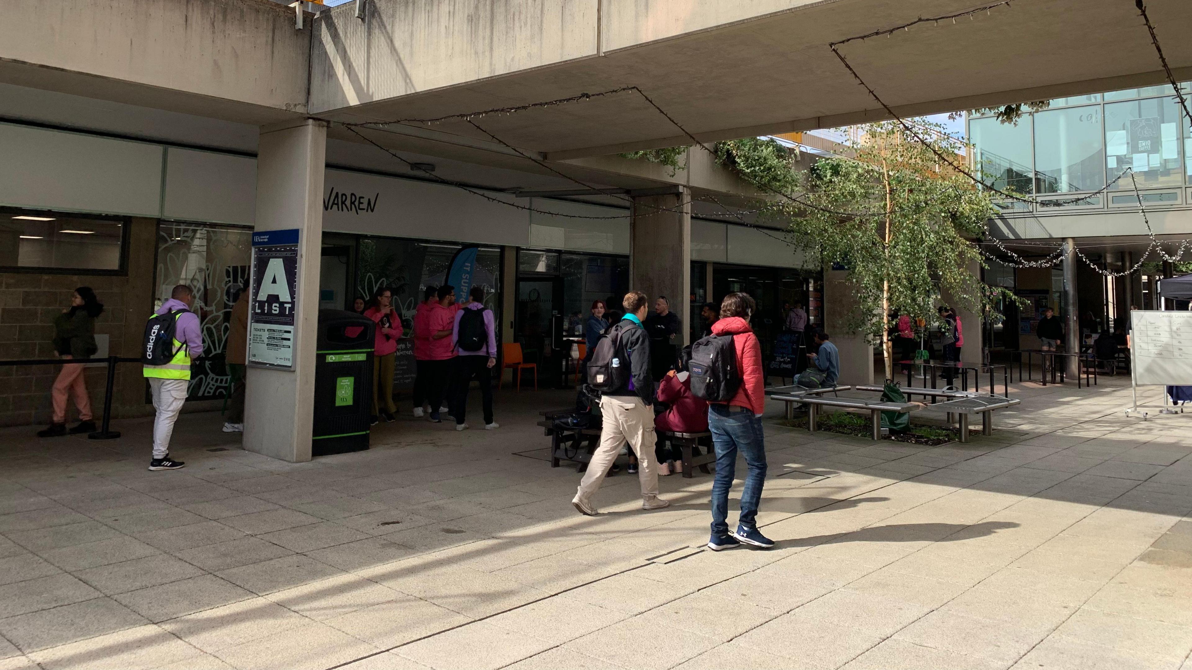 Young university students mill about the concrete built campus. A tree is surrounded by a circular bench and a glass building can be seen to the right of the image.