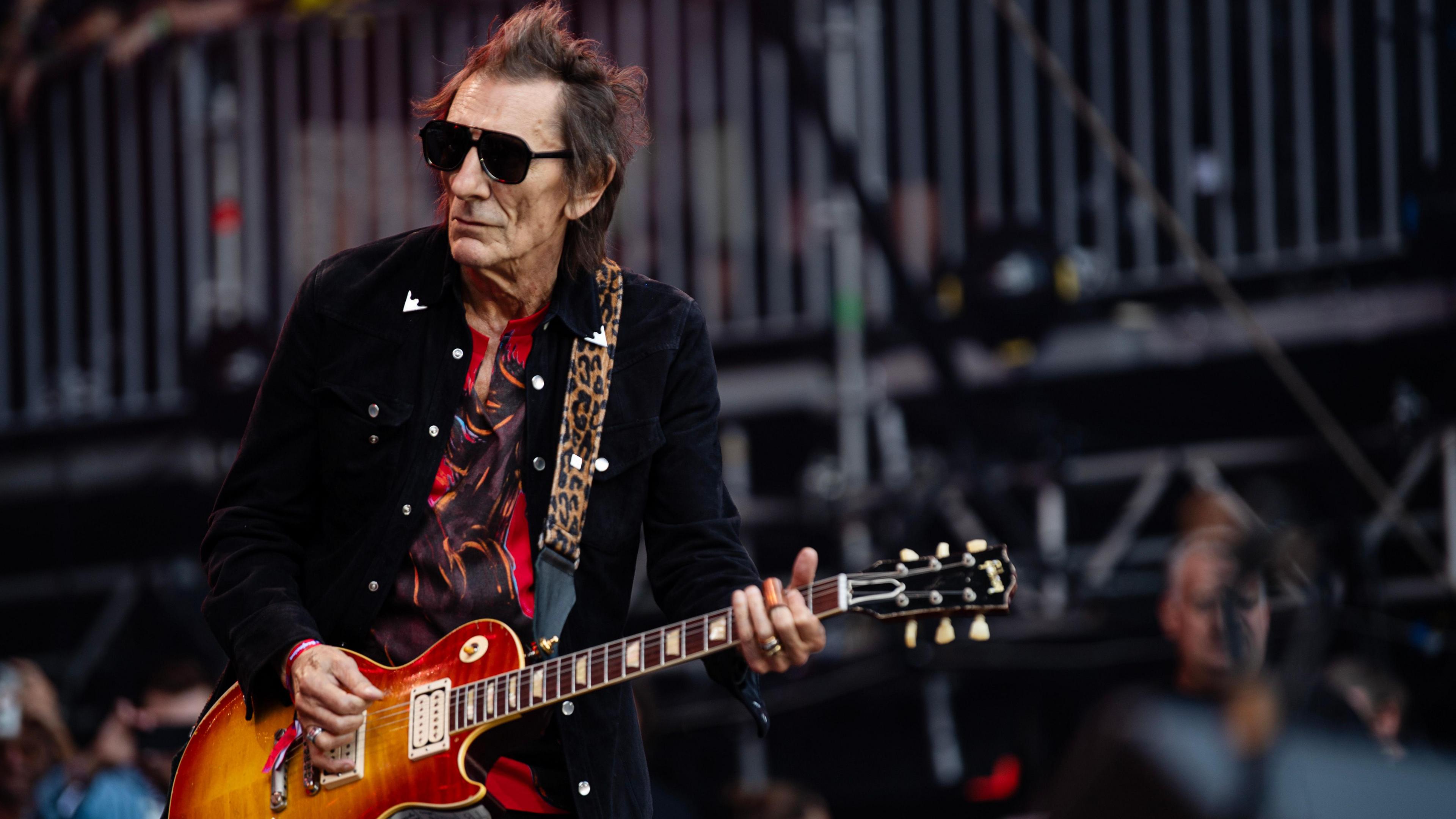A man playing a guitar. He has brown hair and he is wearing sunglasses. He is wearing  red T-shirt and a black jacket.