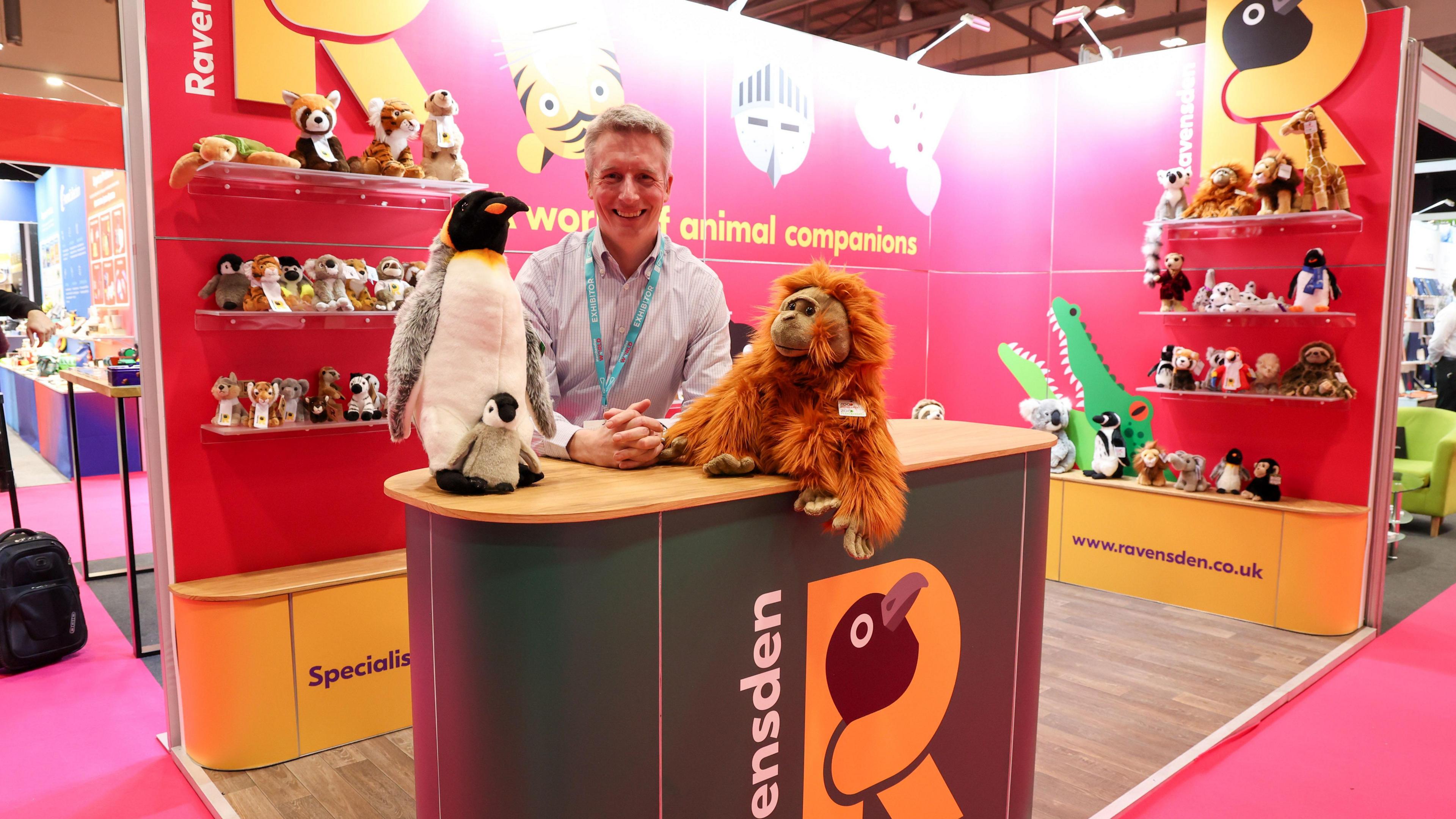 Man in shirt standing at a desk with two penguin toys and an orangutan teddy