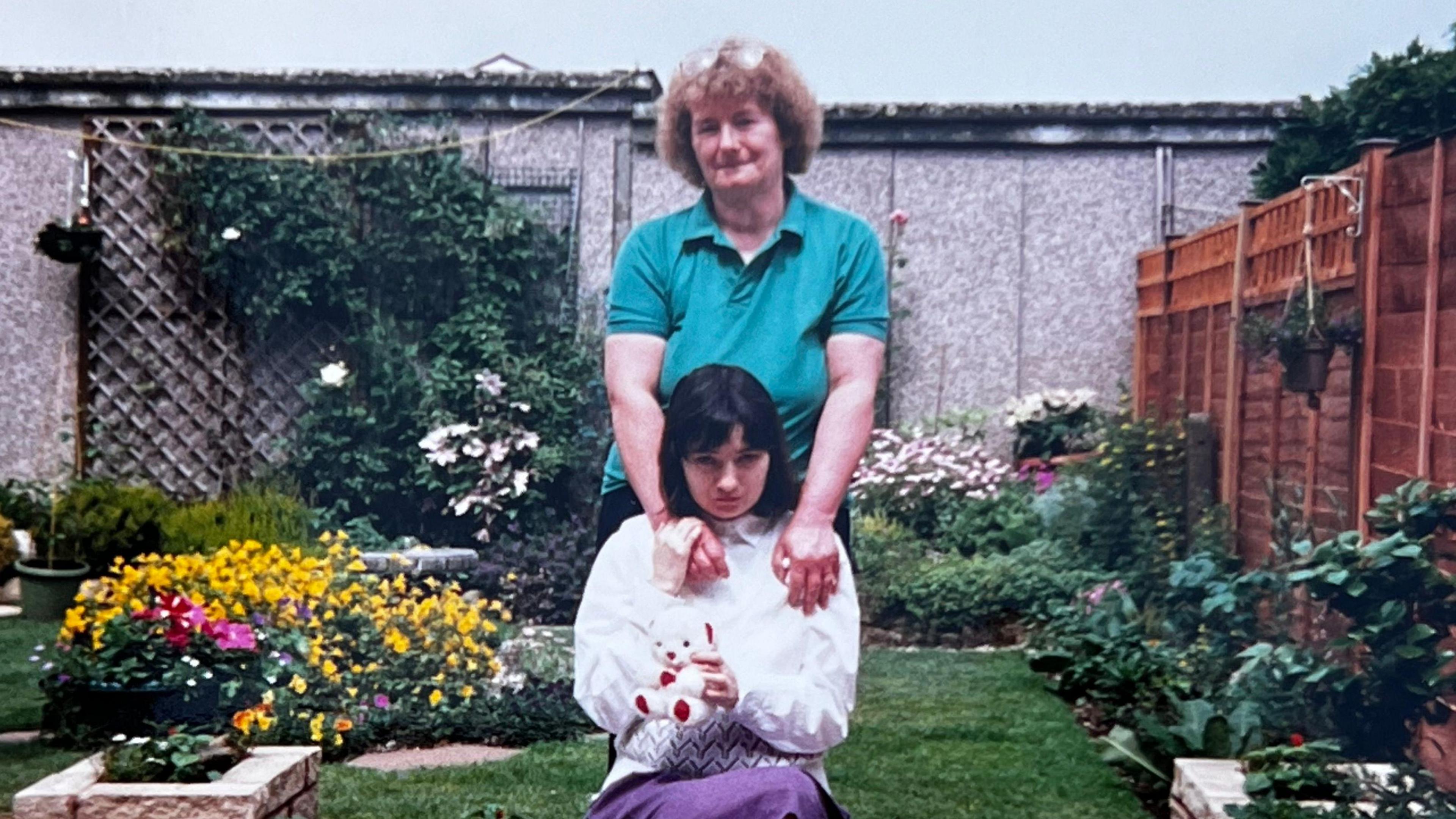 A photo of a girl with shoulder-length black hair, wearing a white shirt and holding a white teddy bear with red features, sits in a garden. Her mother stands behind her with her hands on her daughter's shoulders. She has short brown curly hair with her glasses propped up on her head, wearing a green polo shirt. They are both in a garden with plenty of flowers.