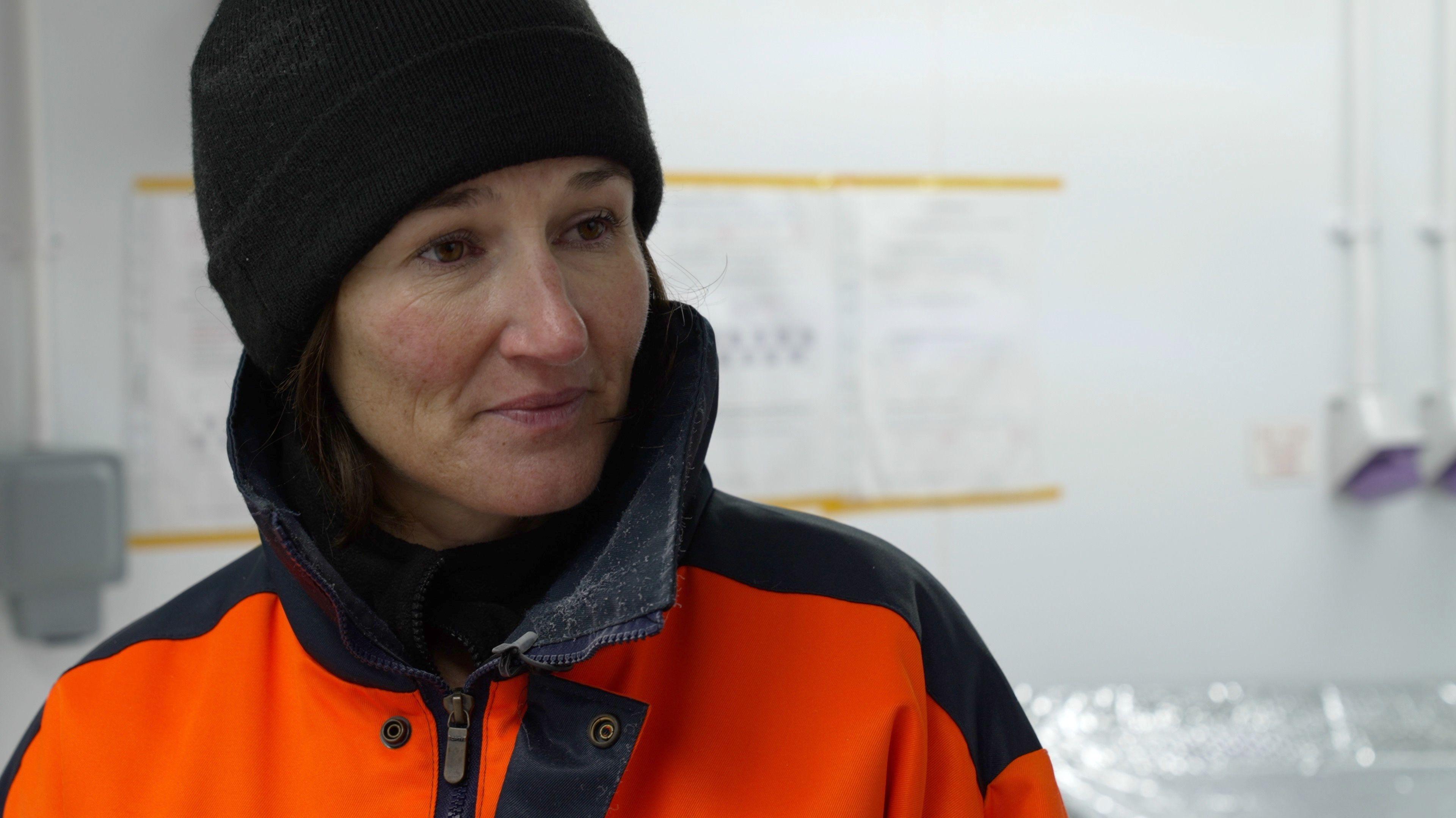 Liz Thomas stands inside a laboratory wearing an orange and black protective suit, and a hat. In the background there is a sign on the wall, and hand sanitiser dispensers.