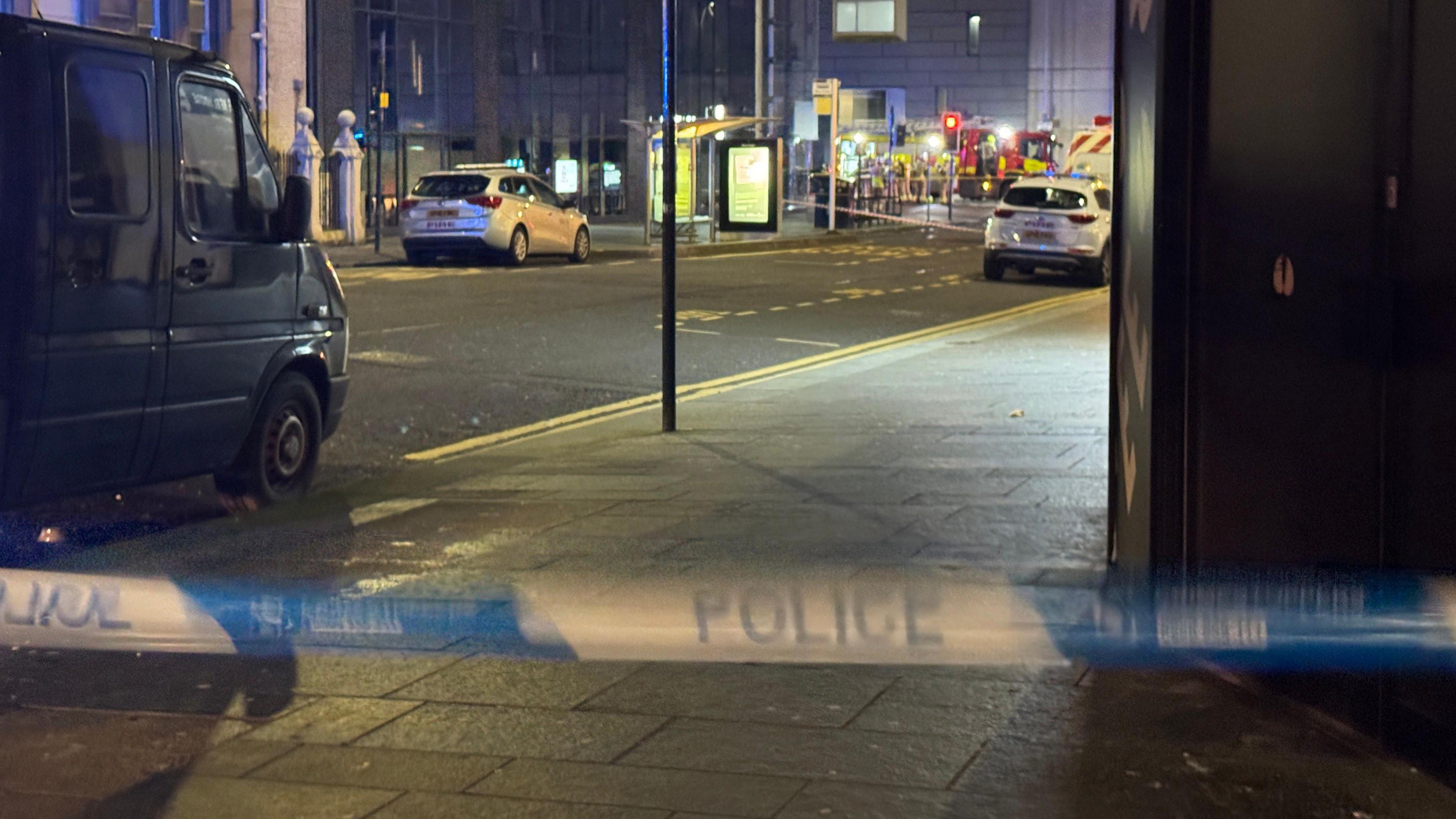 Firefighters can be seen in the distance. A line of blue and white police tape can be seen in the foreground of the image