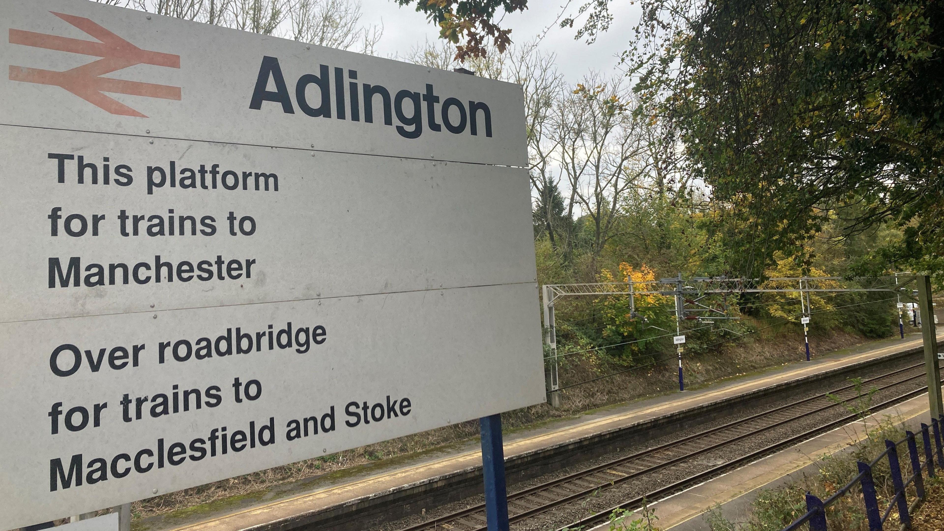 View of Adlington railway station. In the foreground there is a sign saying this platform for trains to Manchester and over the roadbridge for trains to Macclesfield and Stoke. In the background you can see the two platforms.