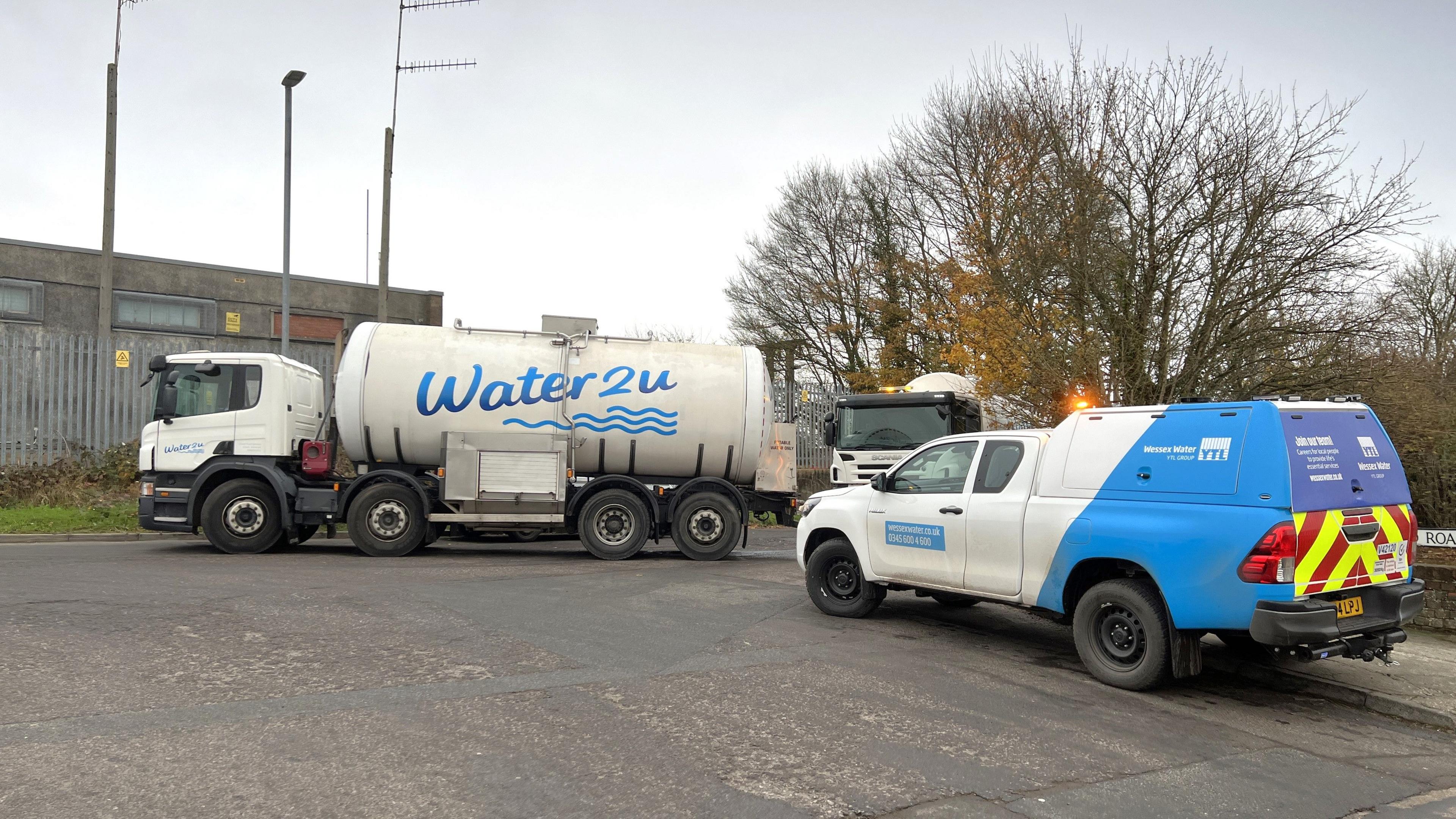 A large tanker truck parked beside the pavement, which reads 'Water 2 U' on the side in blue. Parked behind it is a Wessex Water blue and white work truck. In the background there is an industrial looking building, a spiked metal fence and trees.