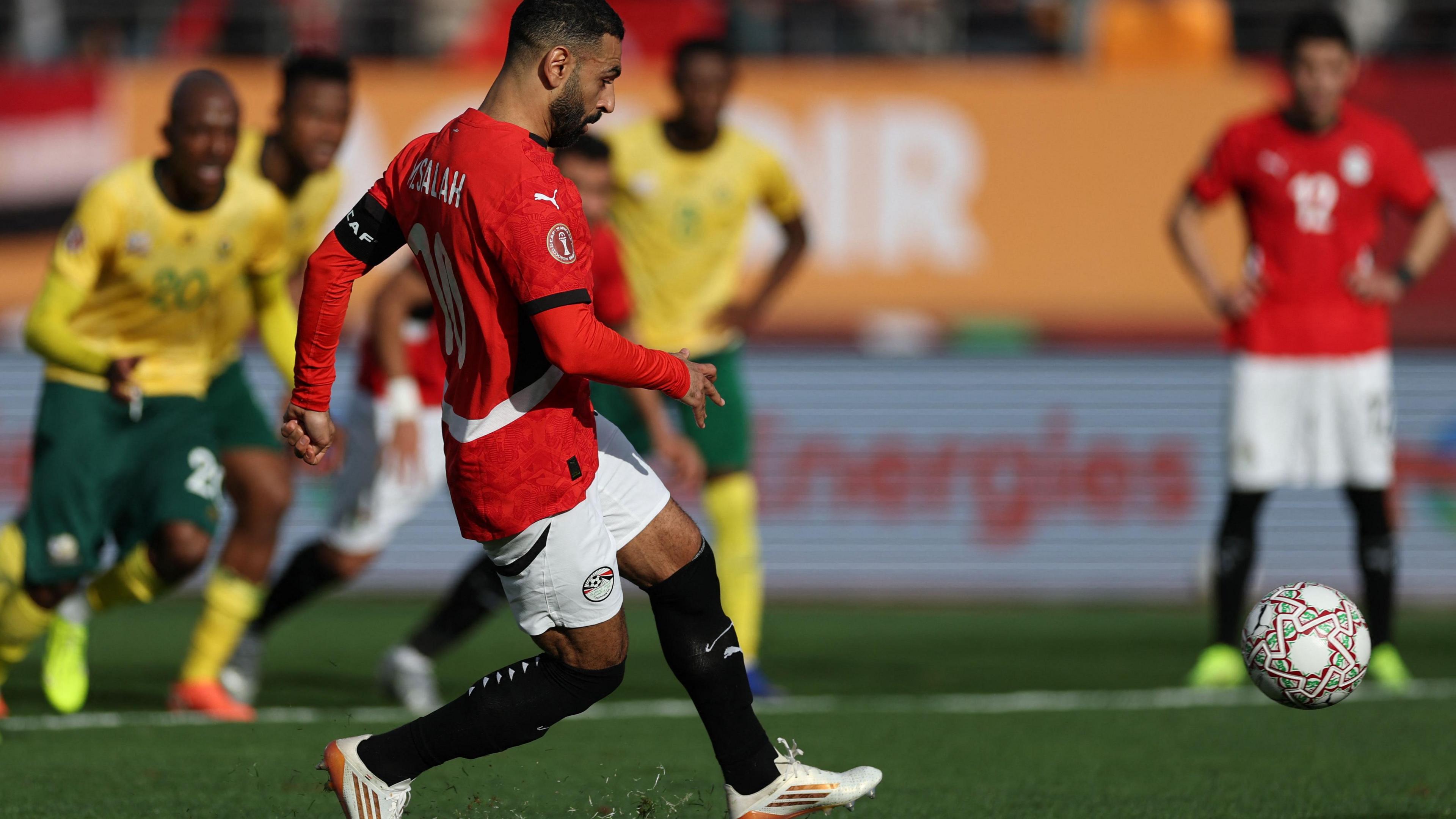 Mohamed Salah, wearing a red Egypt jersey, white shorts and black socks, shoots with his left foot and scores a penalty kick against South Africa at Afcon 2025.