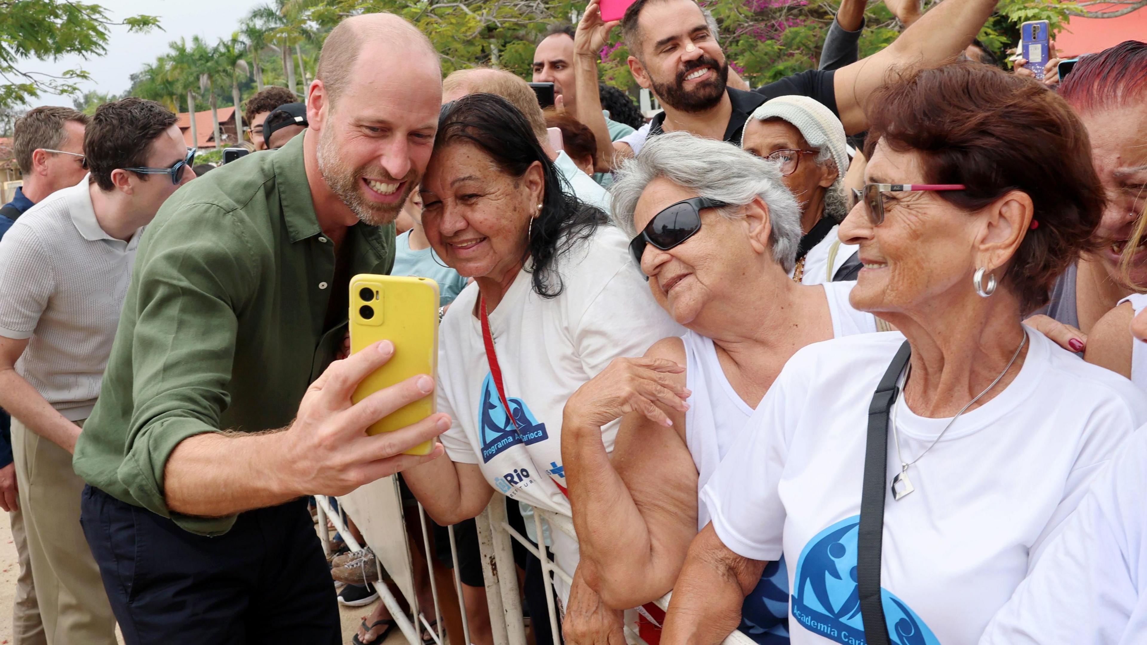 William stands on the left and poses with a woman for a selfie, which he takes holding her phone in a pastel yellow case. There are two other women next to her on the right of the picture, and behind them are the crowds
