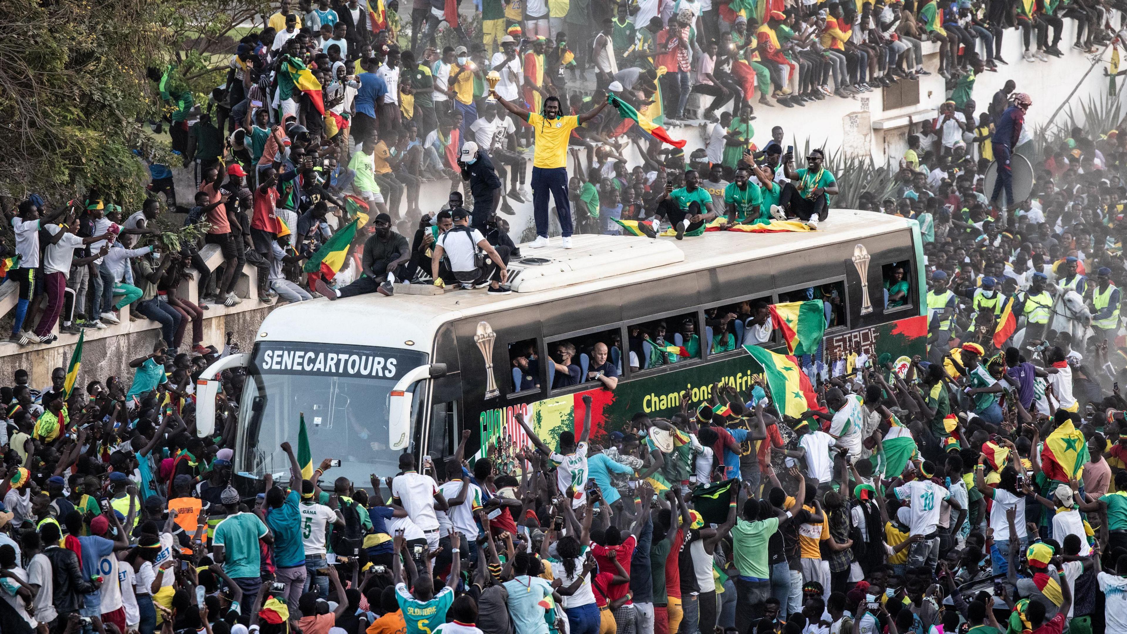 A large crowd, wit several waving Senegalese flags, crowd around a bus carrying the national squad as several players sit on top of the vehicle and Aliou Cisse stands aloft wearing a yellow t-shirt and holding the Africa Cup of Nations trophy