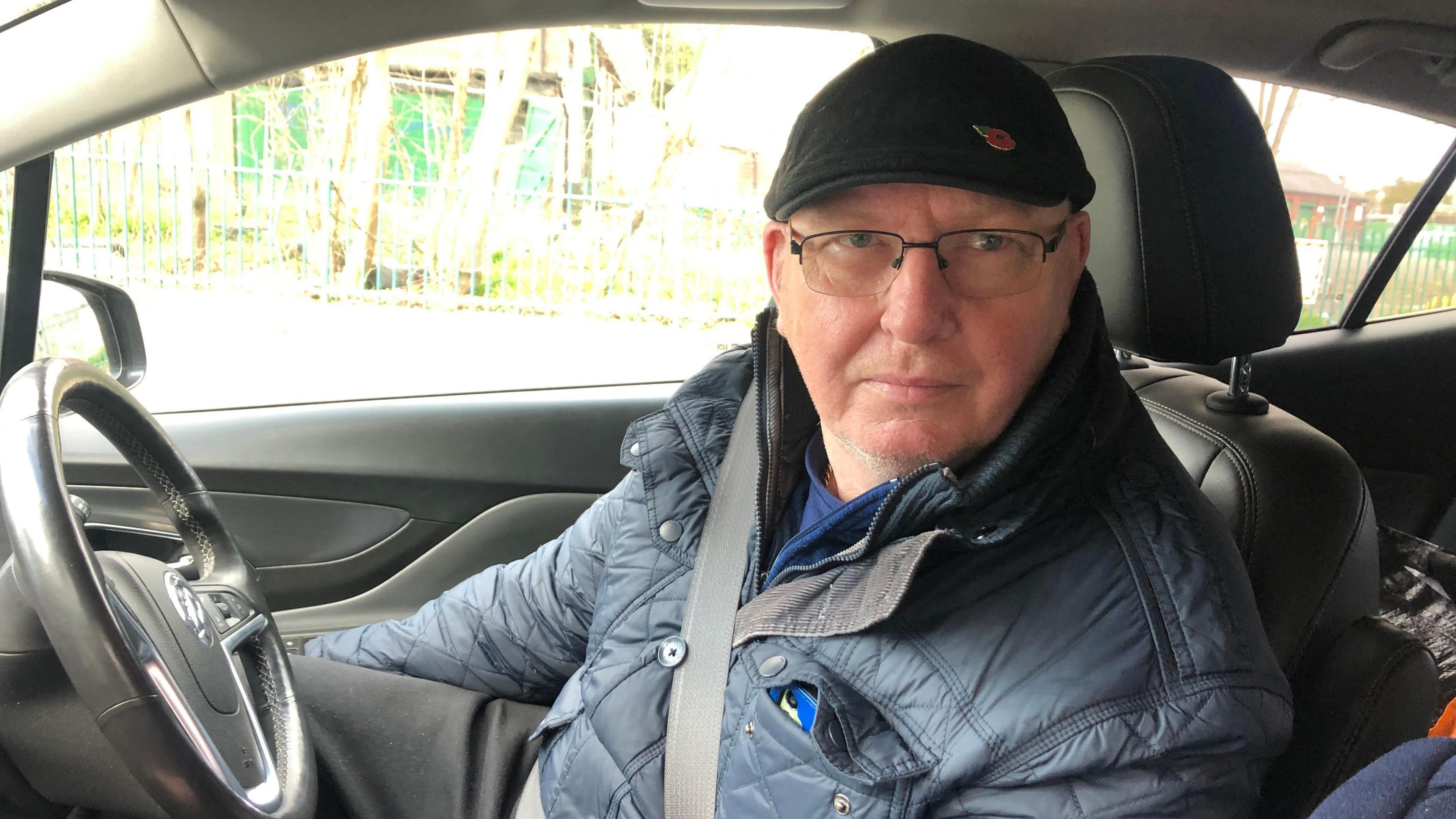 Steven Reeves in a car. He is next to a steering wheel and wearing a seatbelt. He is wearing a farmer-type jacket and a flat cap with a poppy on. 