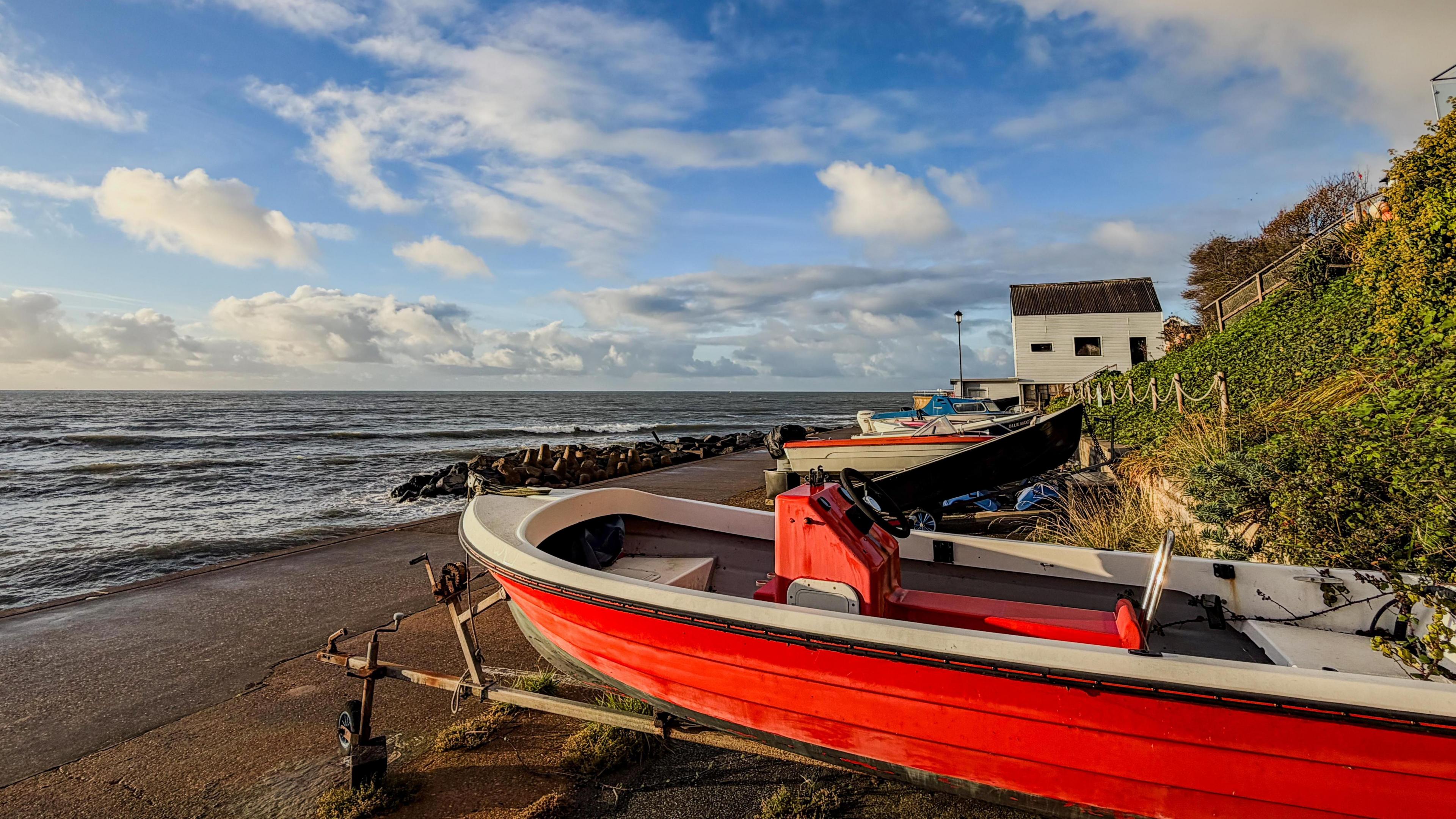 Small boats rest on the shore at Bonchurch, Isle of Wight. The nearest one, on a trailer, is bright red, contrasting with a blue, cloudy sky and green vegetation covering a coastal embankment leading up from the sea.