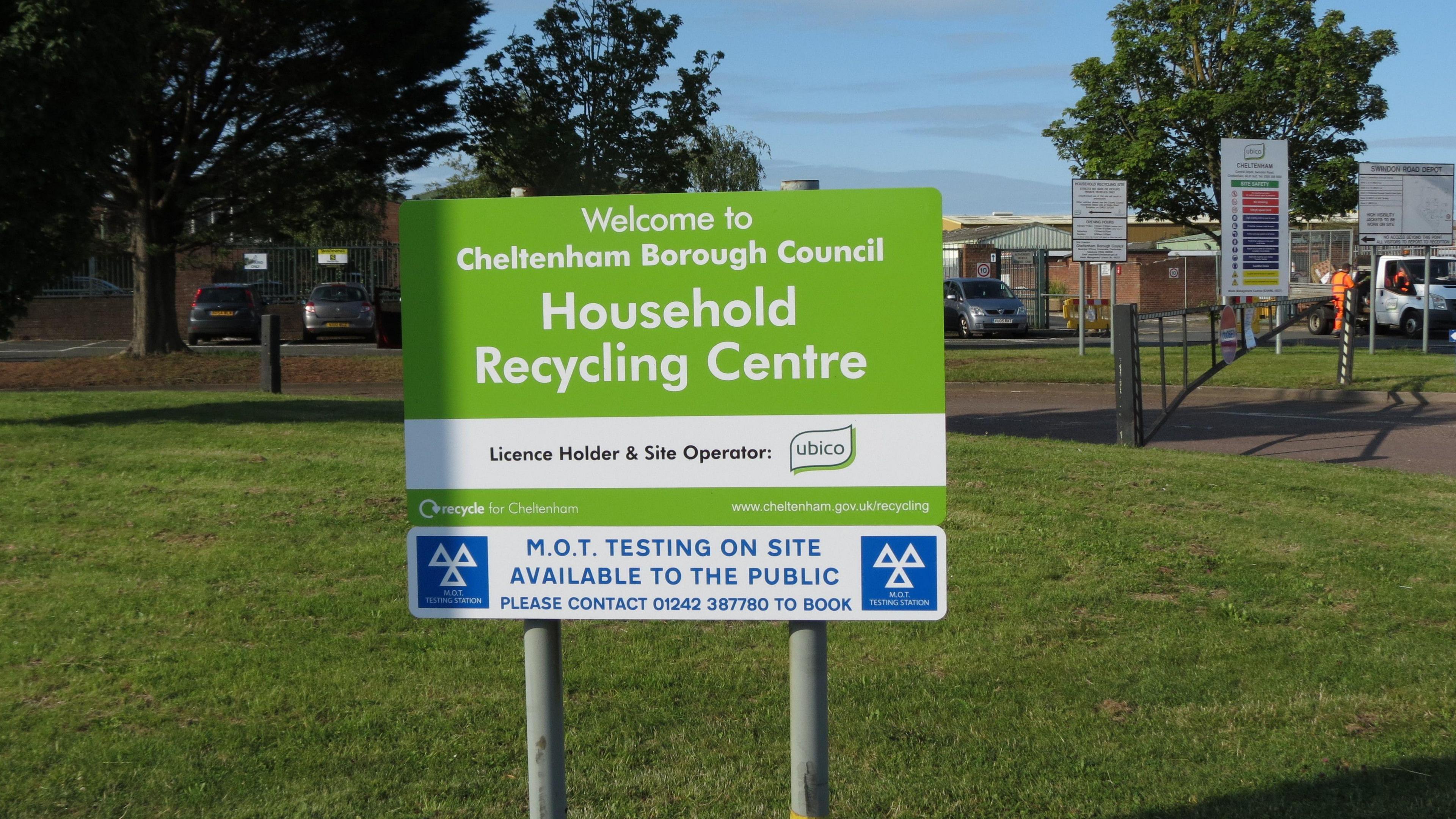 A green sign on two metal posts on a lawn near a recycling centre. It reads "Welcome to Cheltenham Borough Council Household Recycling Centre". The facility can be seen in the background, as can a car park and trees.