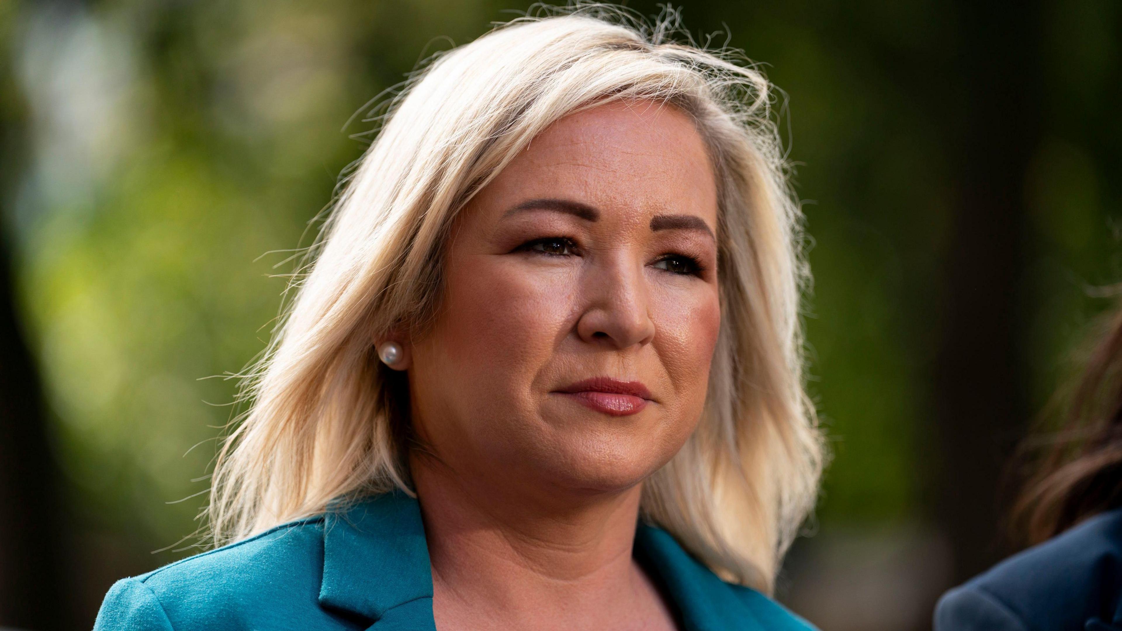 Michelle O'Neill is wearing a blue suit. She's talking to the media. She has shoulder length blonde hair and is wearing pearl earrings.