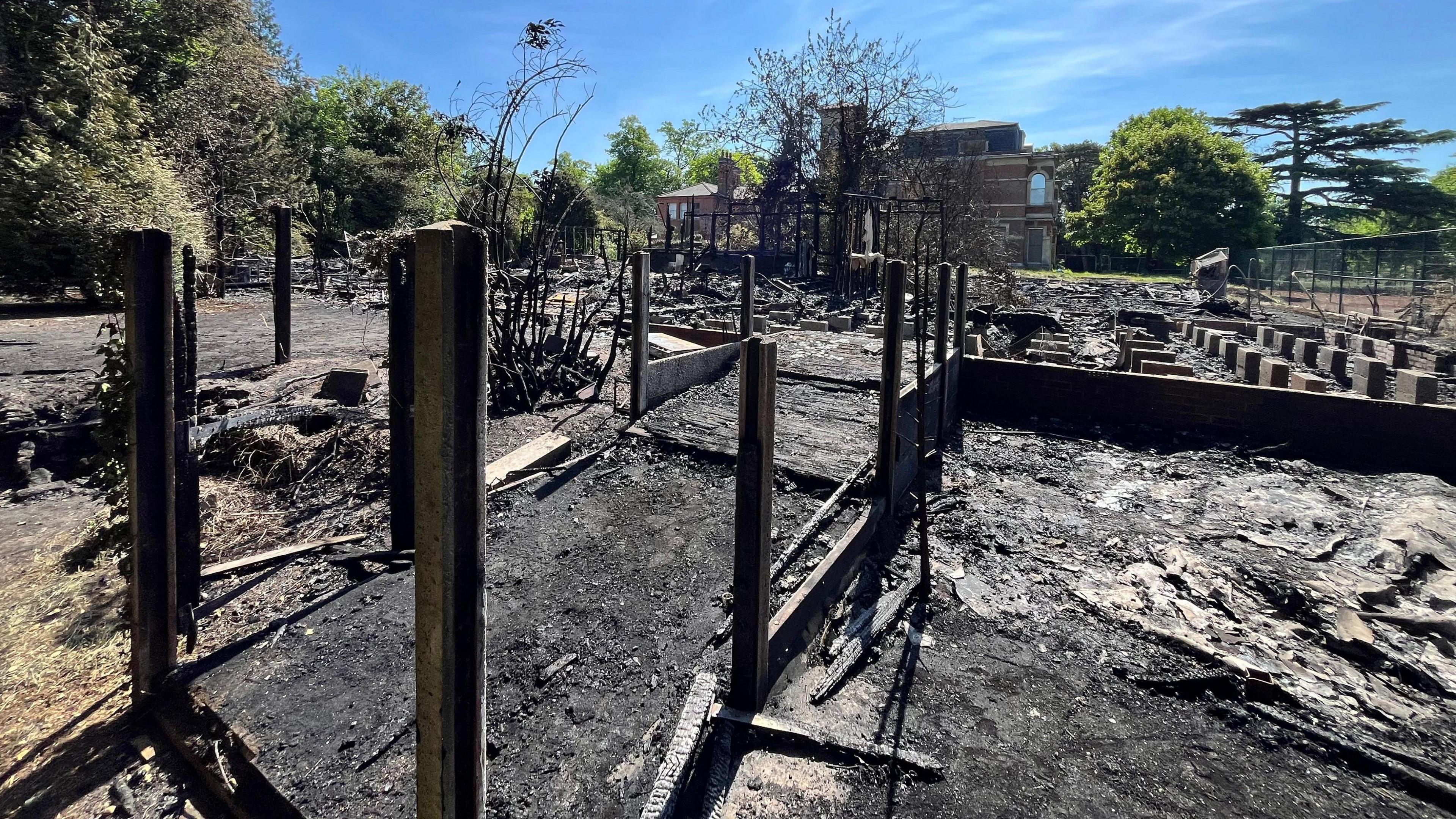 The remains of a burned out building on a sunny day. There are no visible walls. The ground is charred black.