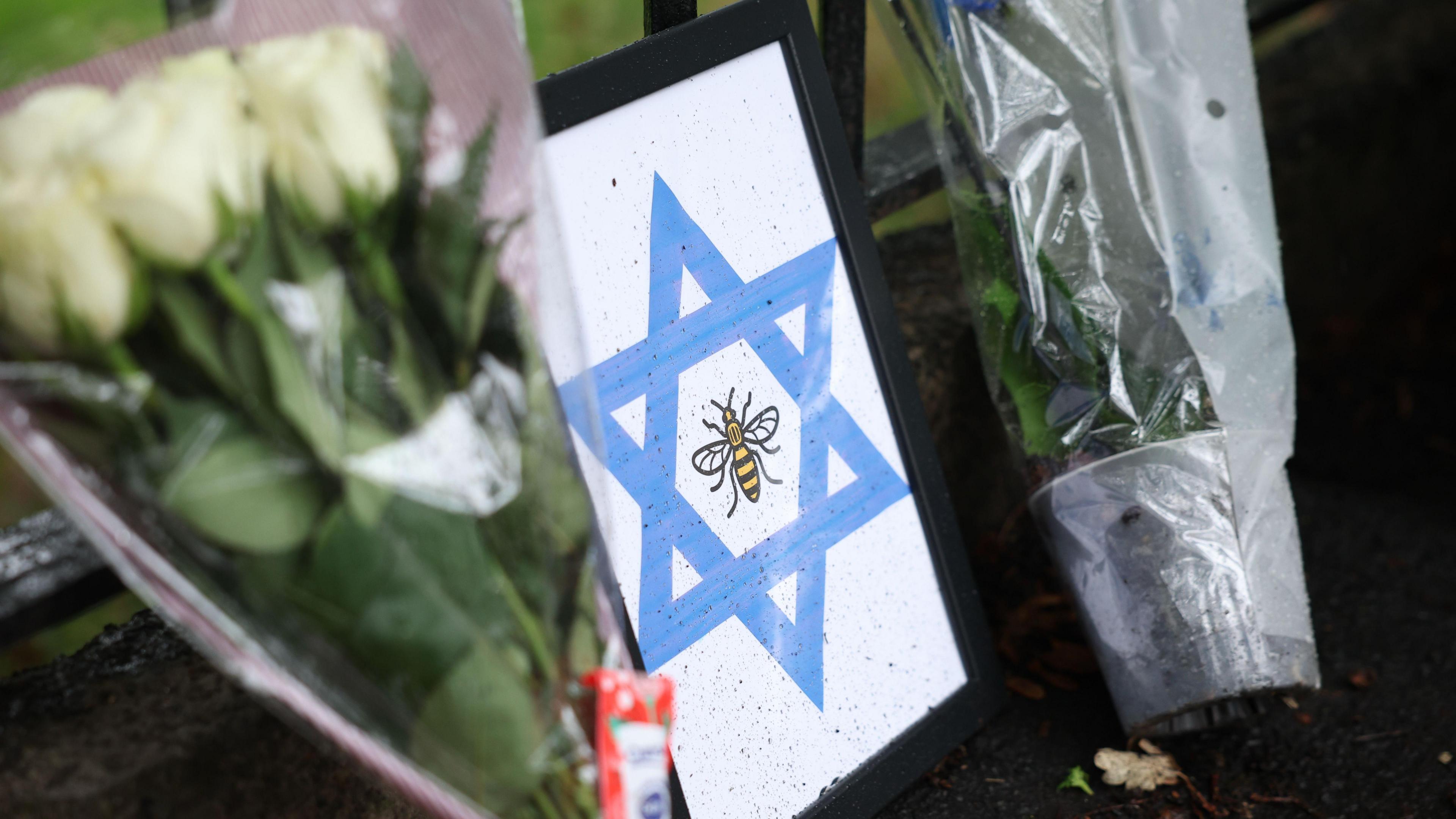 Bouquets of flowers are leaning on a black fence near the synagogue. They are either side of a blue Star of David printed on white paper and framed. The Manchester bee is in the centre of the picture.
