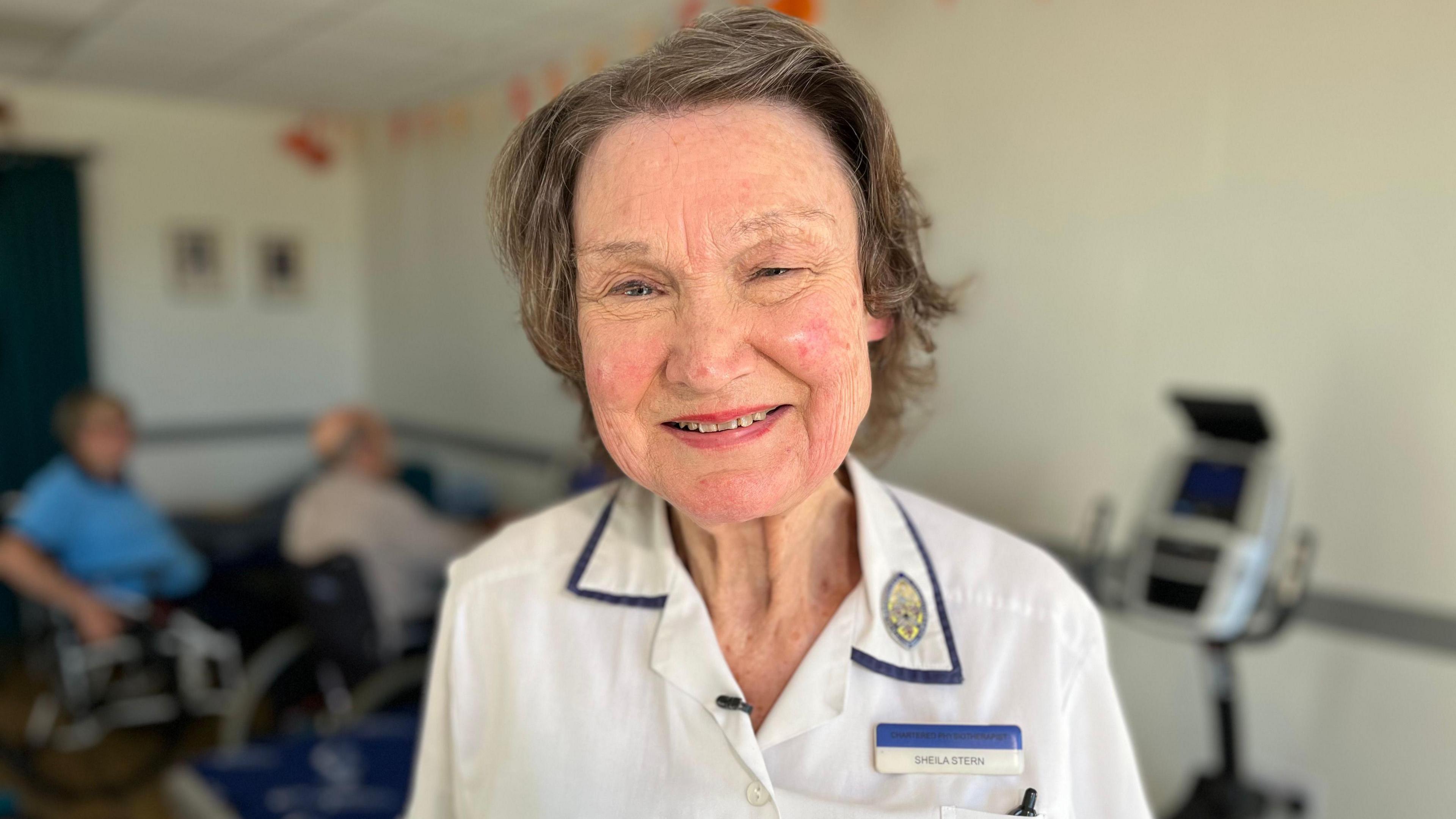 Headshot of Shelia Stern smiling at the camera while at her weekly physio class. She wears a white medical uniform with blue around the edges of the collar and a logo on the right collar. She also has a name badge above her her top right pocket, which contains a pen. The background is blurred but two figures, who look to be using wheelchairs, can be made out participating in the weekly class. 