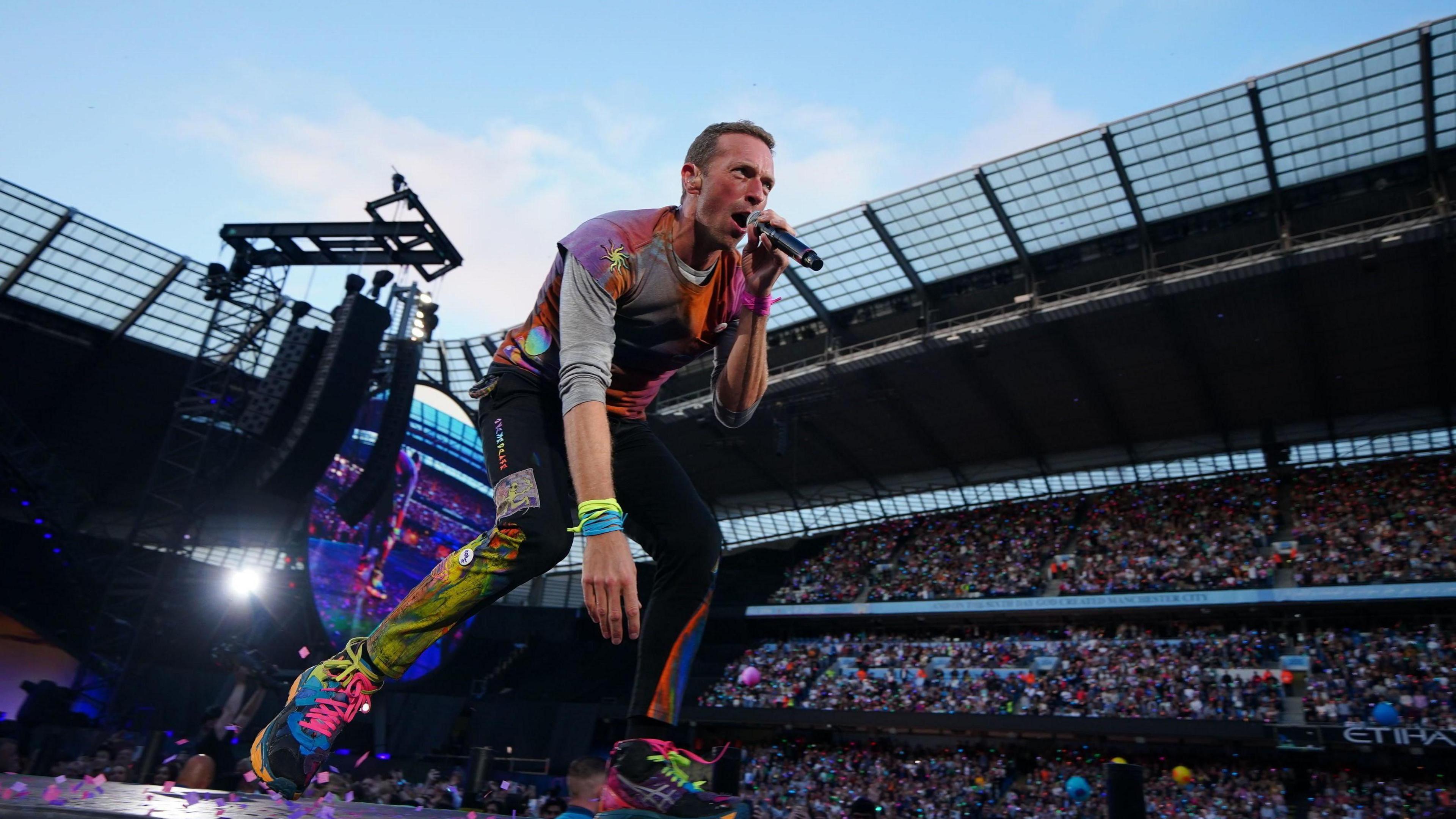 Chris Martin, wearing paint coloured clothes, performs on stage at the Etihad Stadium in Manchester.