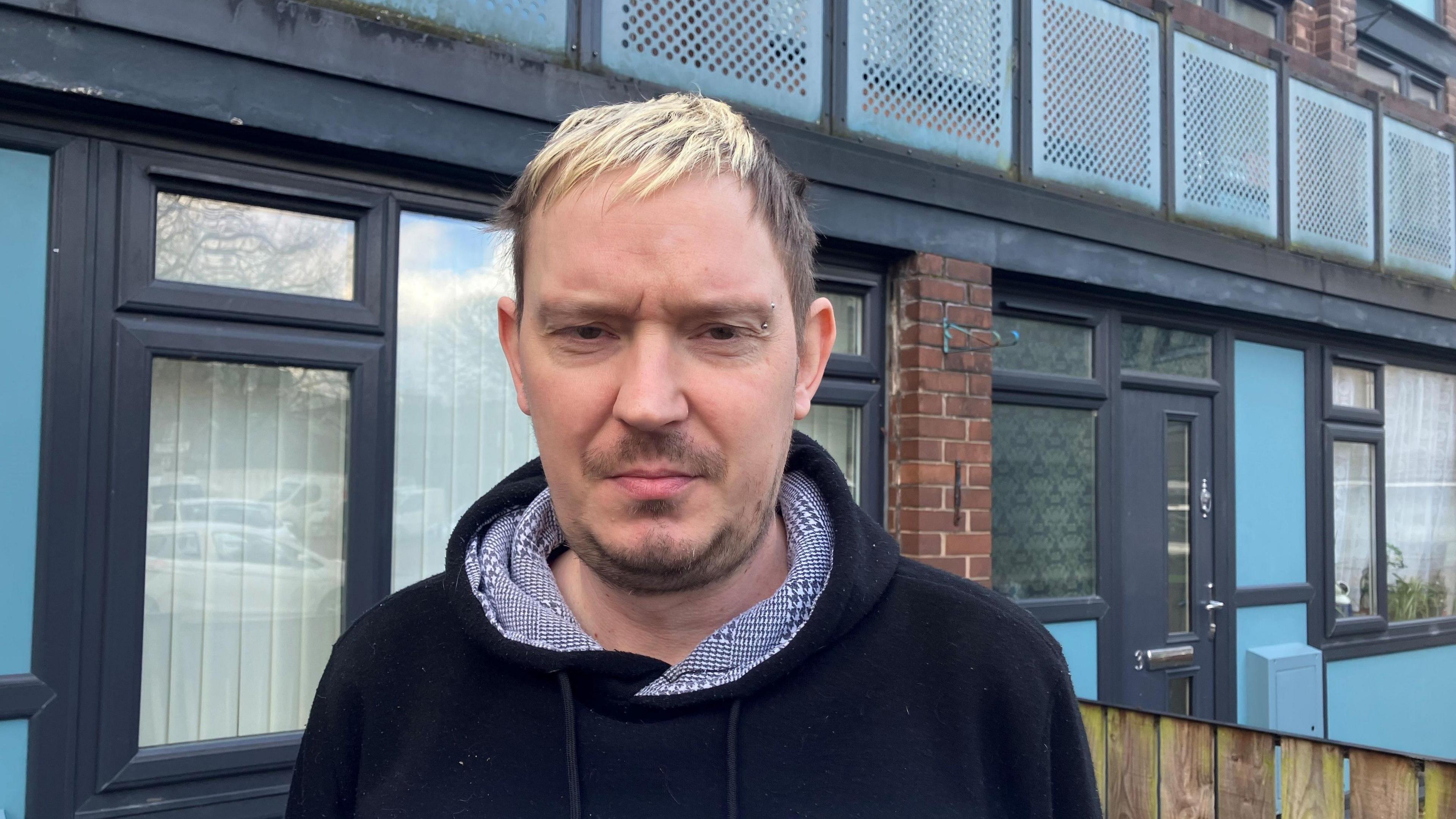 Photograph of Barry Smith outside his home on the Mottram Street estate in Stockport. Barry has short bleached blond and brown hair, a goatee and a pierced eyebrow, He is wearing a black-hooded top with a houndstooth lining.