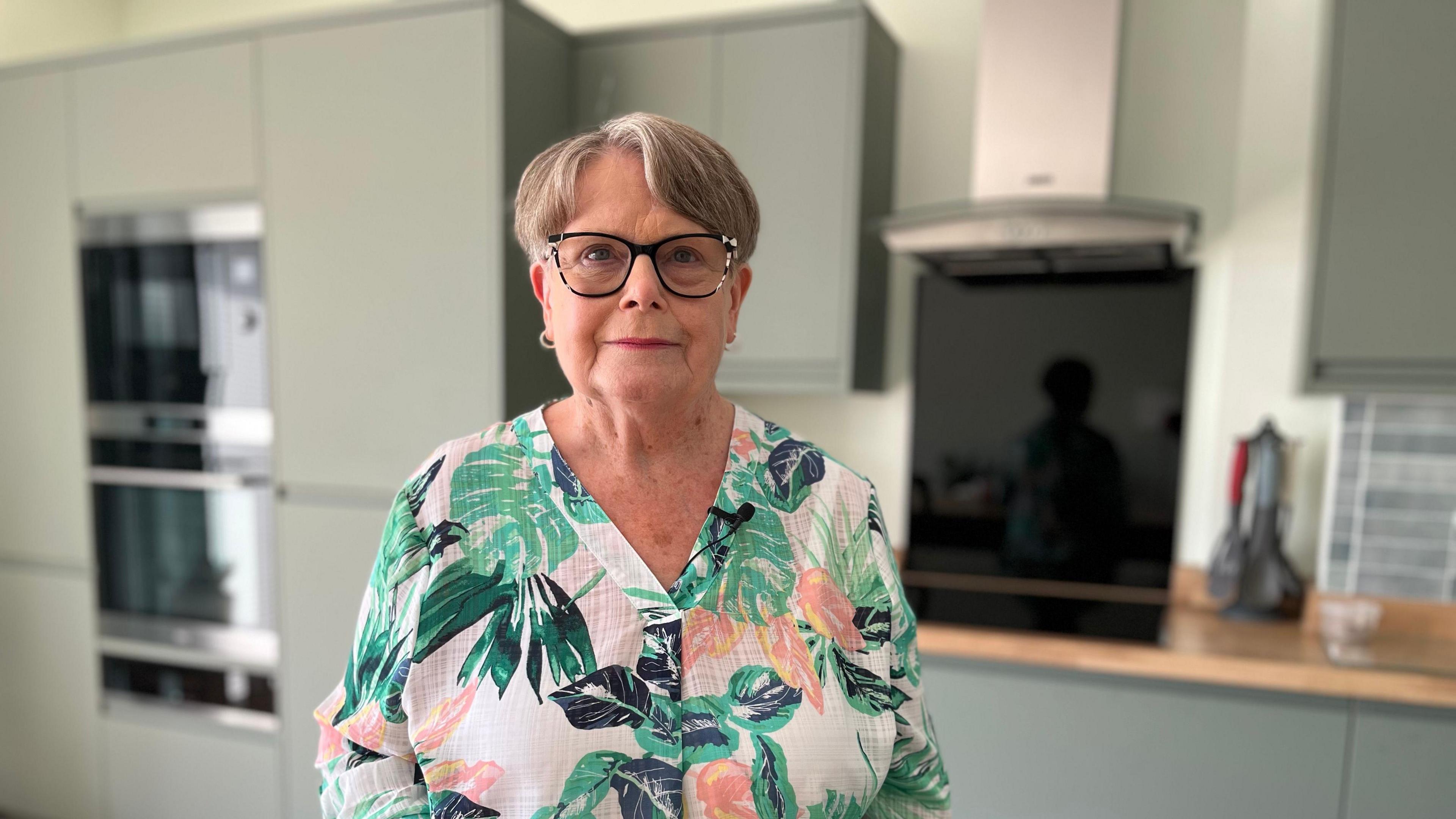 Carole Hager stands in the kitchen of her newly rebuilt house. She has short grey hair and is wearing black-rimmed glasses and a brightly coloured floral shirt. Behind her is a modern kitchen with sage green kitchen cupboards and wooden work surfaces.  