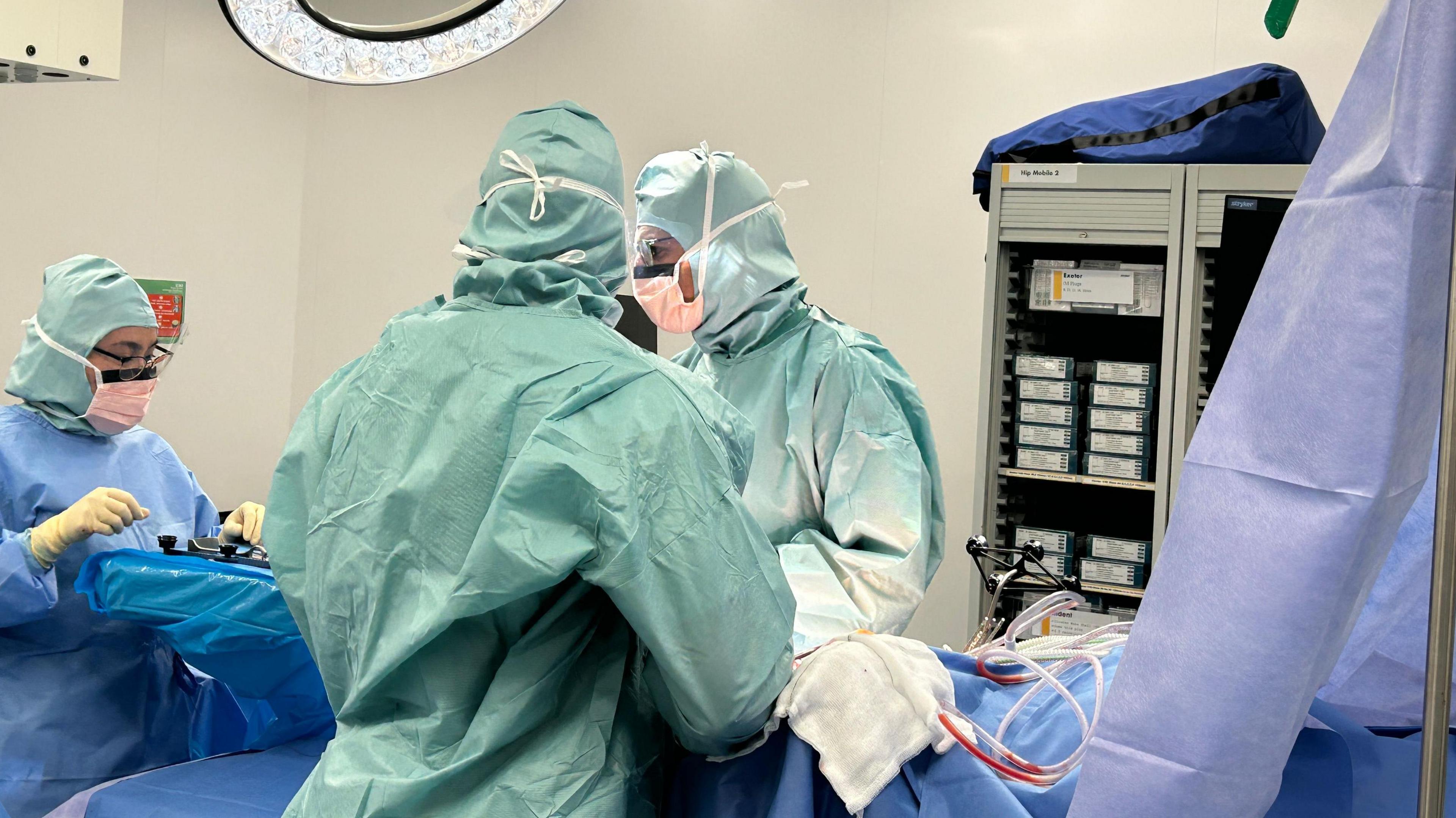 Three people wearing green scrubs stand over a surgical table. There is a person on the table who cannot be seen in the image. They are performing hip surgery. 