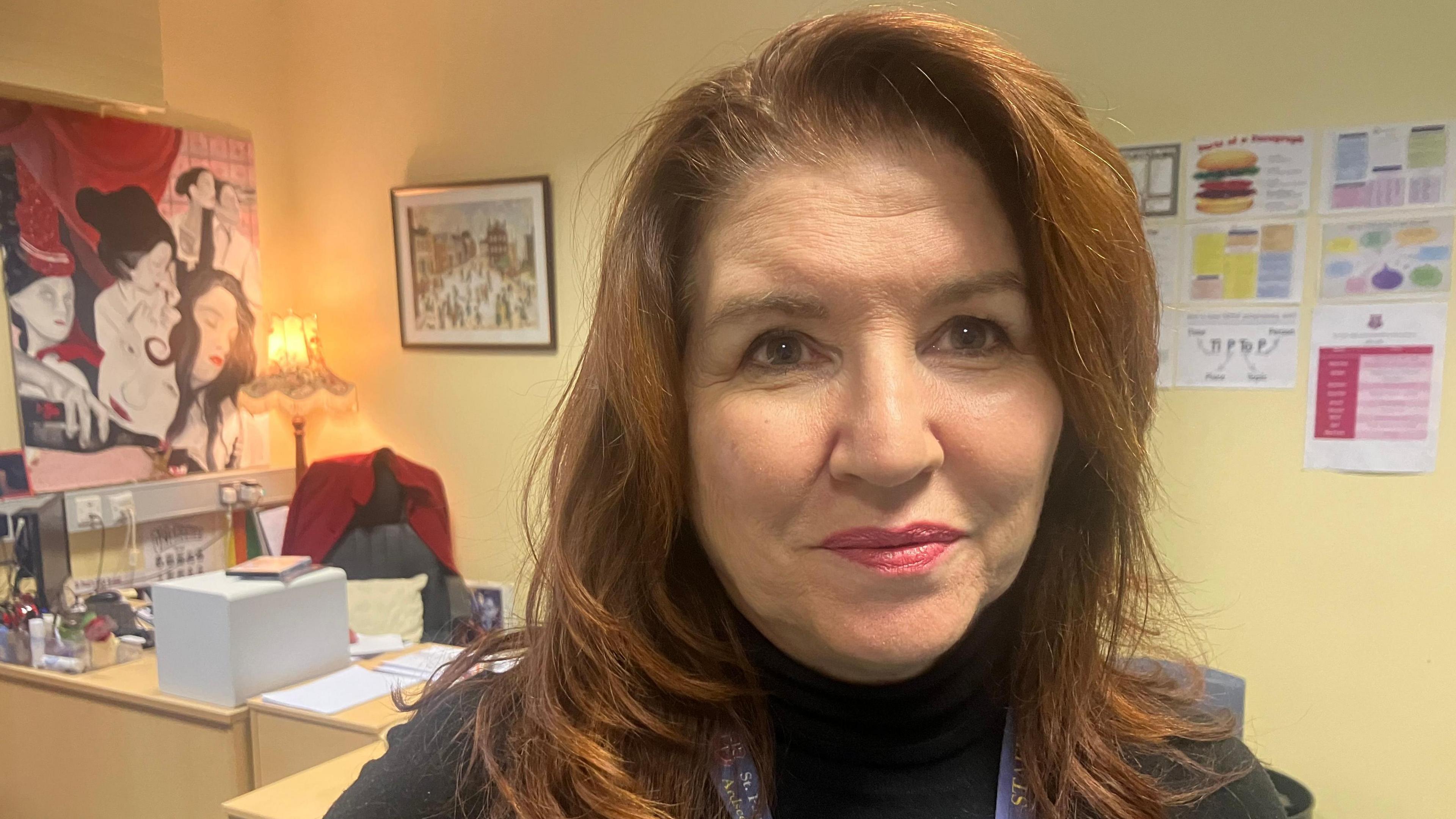 Pauline Lynch with long ginger hair in a close-up shot staring at the camera. She wears a black top and has pink lipstick on. In the background is an office chair and desk with posters and a print on the wall.