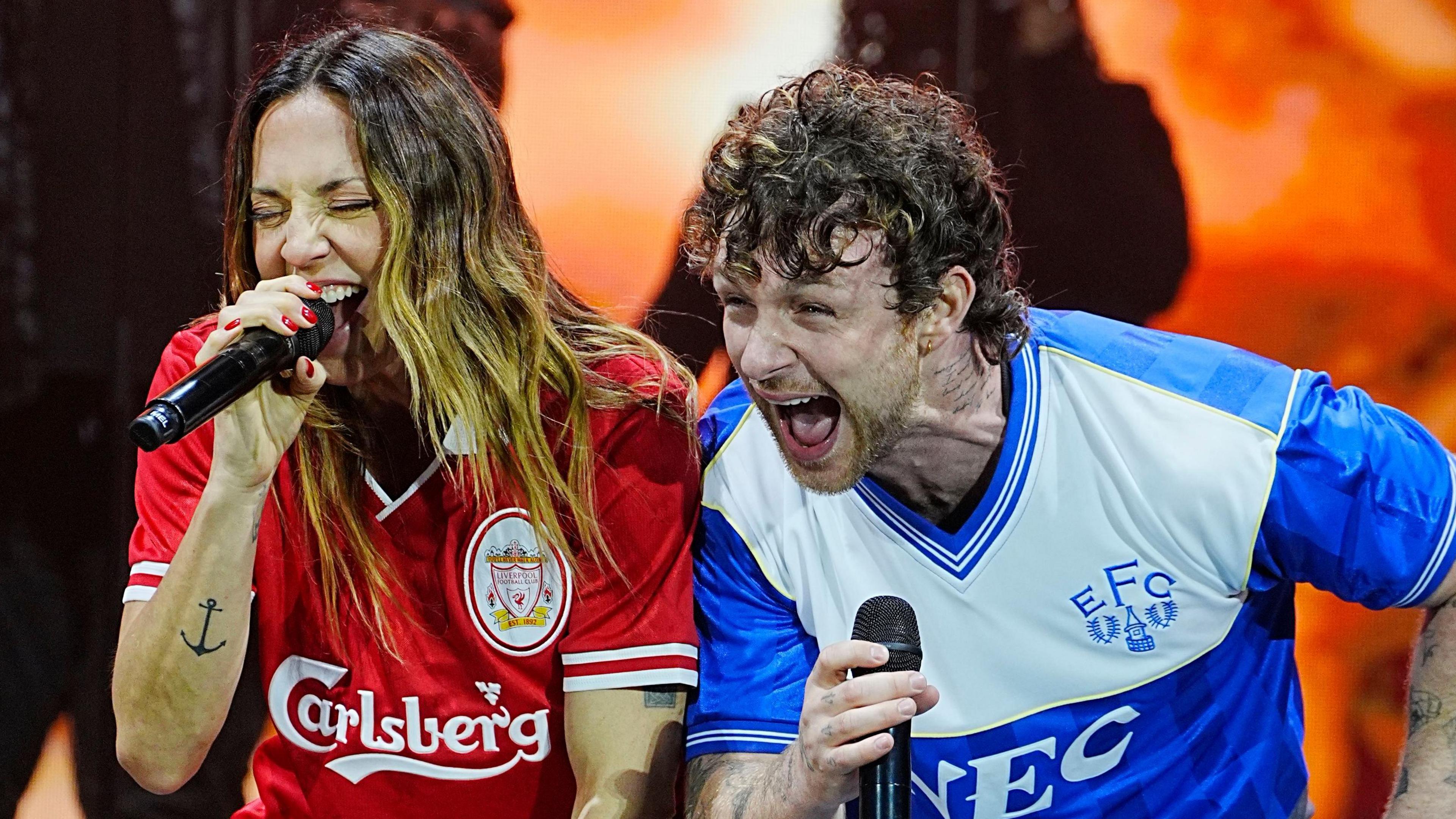 Mel C (left) wearing a Liverpool football club jersey holds a microphone to her mouth as she belts out a song with Tom Grennan, dressed in an Everton jersey
