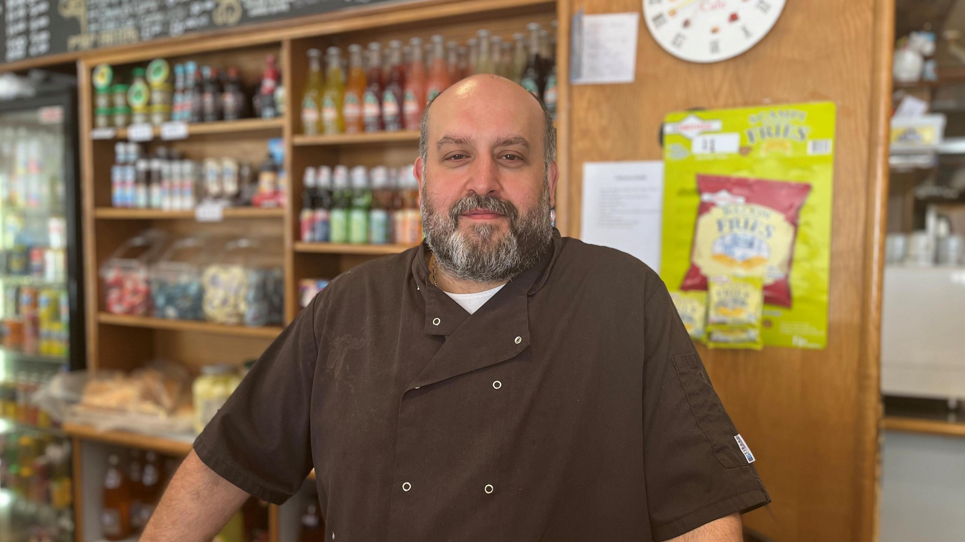 Adriano Pia with a shaved head and a full beard wearing a dark chef-style jacket, standing behind a counter in a shop with shelves of bottled drinks and snacks, a wall clock, and a “Savoury Treats” display visible in the background.