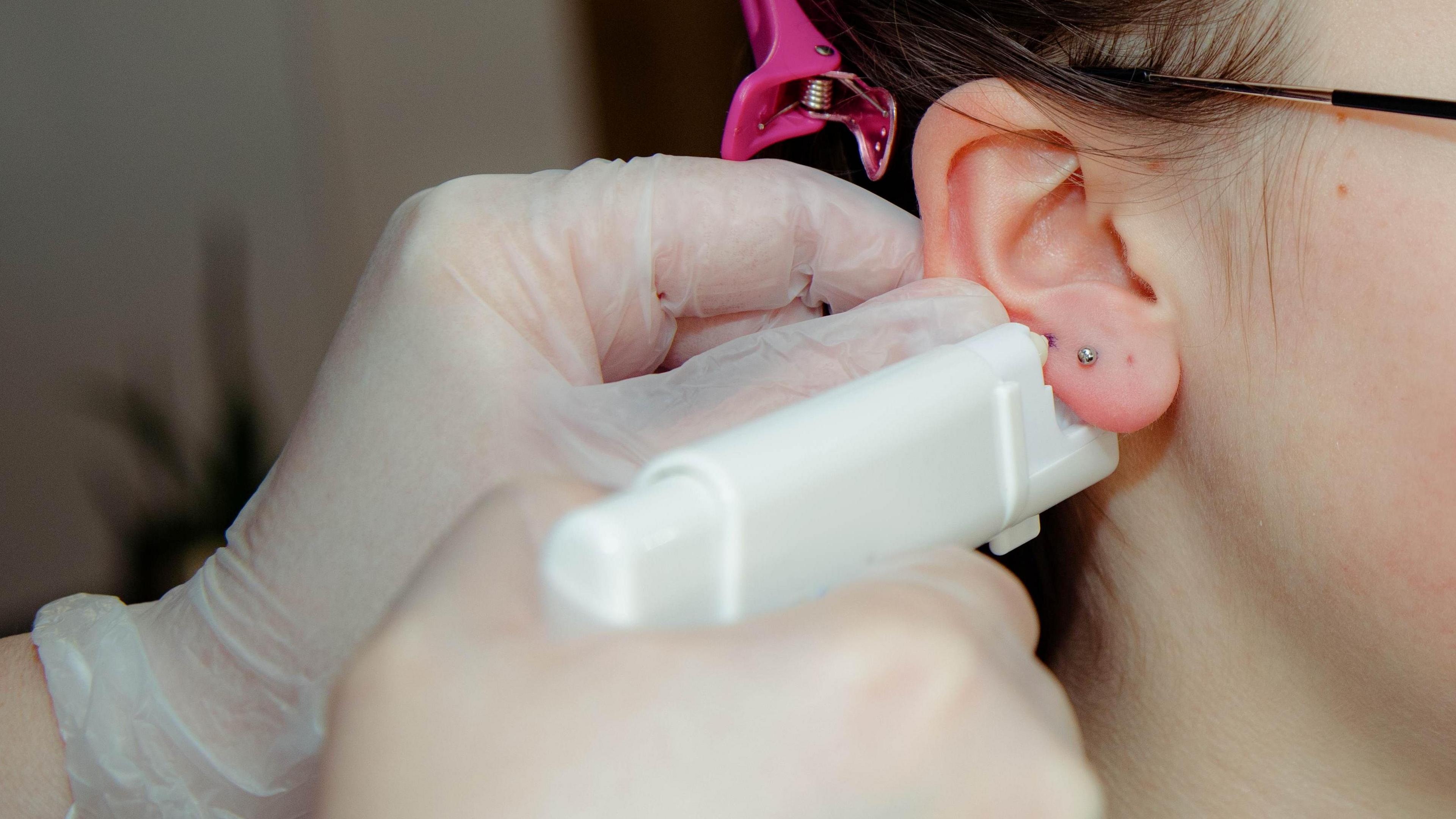 Close up of a hand of unrecognizable adult in protective gloves holding piercing gun in hand and preparing to make puncturing in earlobe of a teenage girl