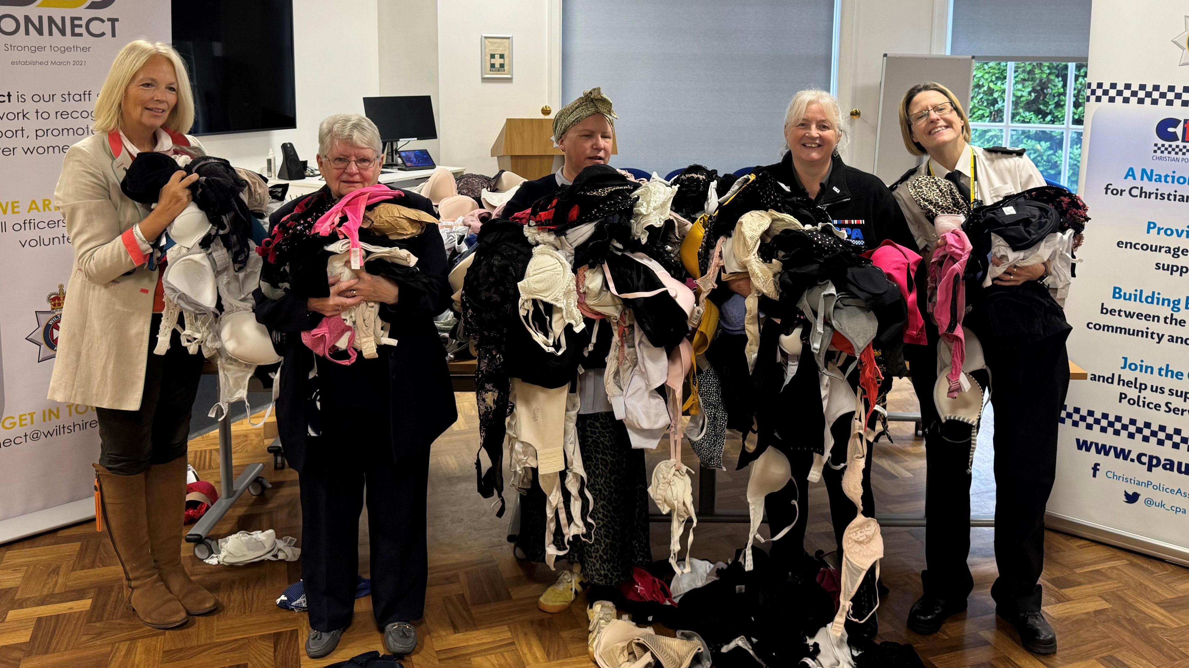Five women hold huge piles of bras, of all shapes and sizes, which are spilling onto the floor as there are so many.