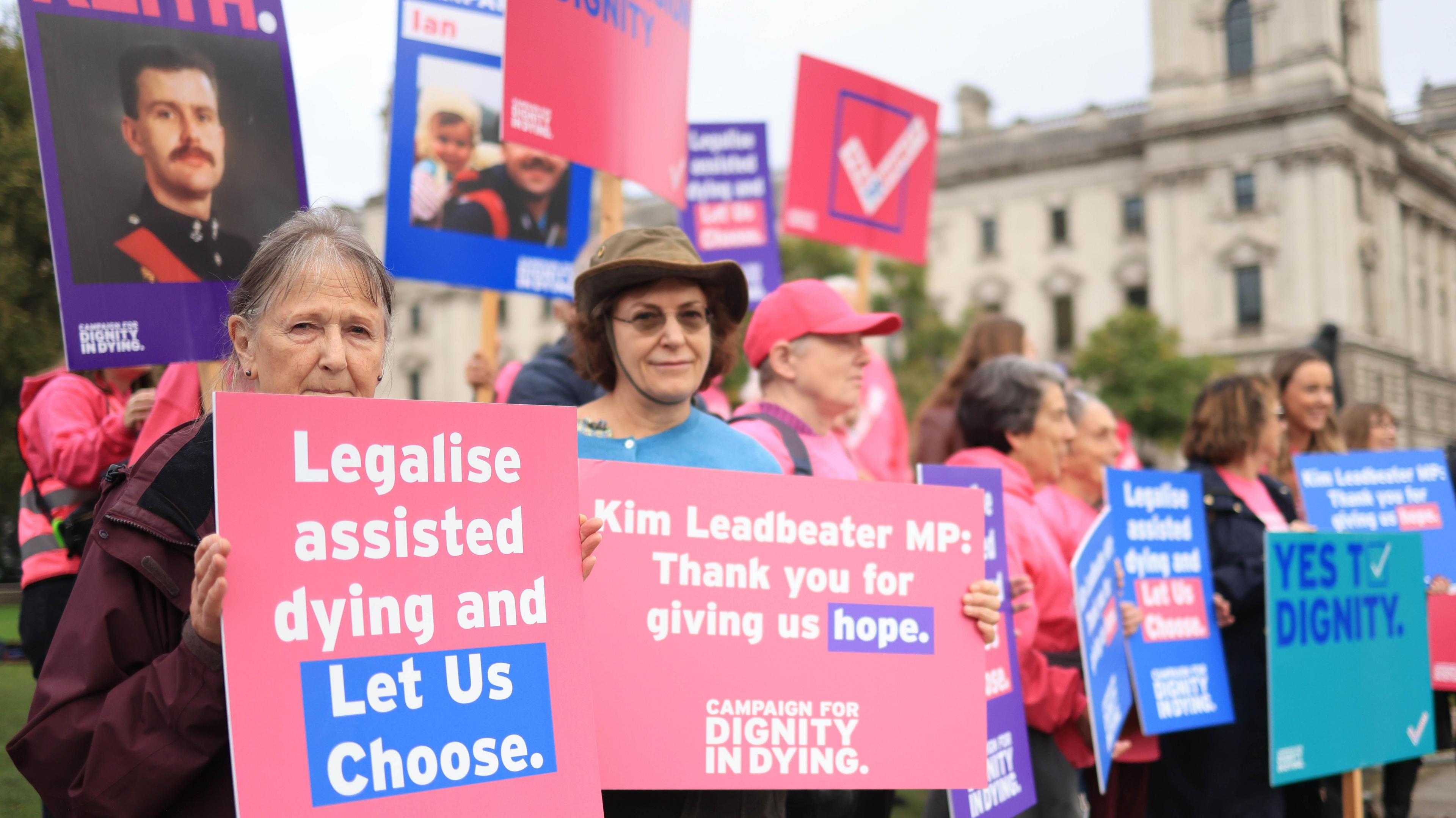 Supporters of the Terminally Ill bill stand in front of Parliament. They are holding up pink and blue signs in support of the bill. 