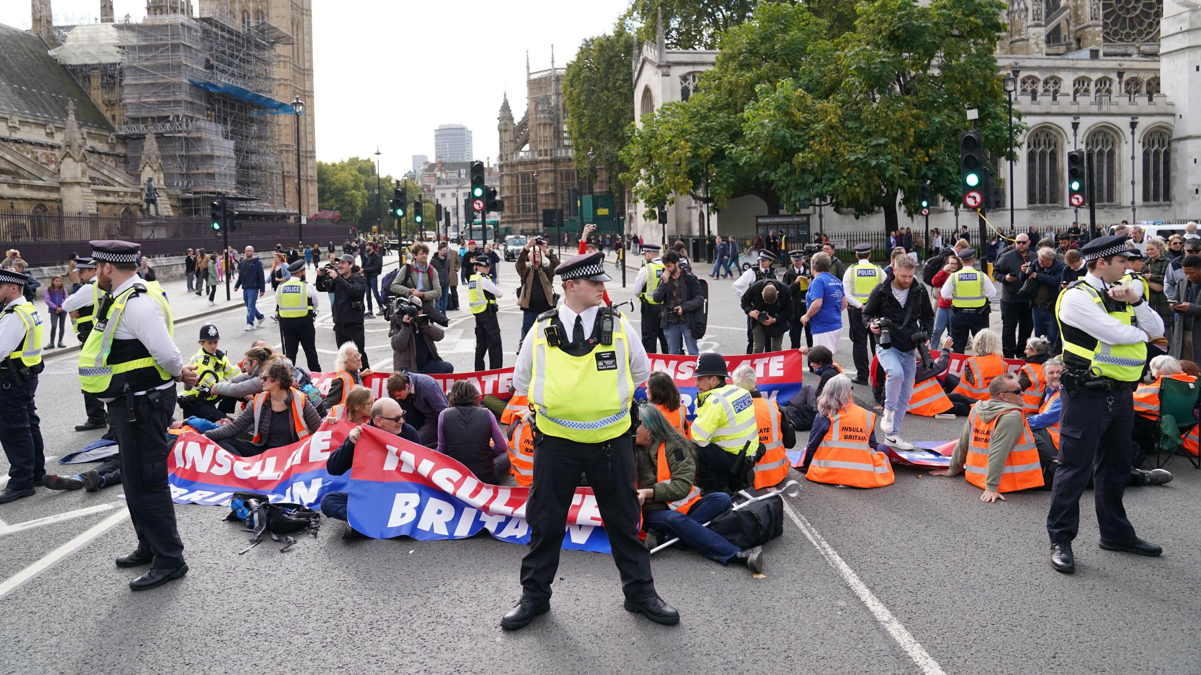 Insulate Britain protestors sat in the middle of a road in London. Many of them are wearing orange high vis jackets. They are holding banners with the Insulate Britain logo on it in white letters. Police officers are standing around the group.