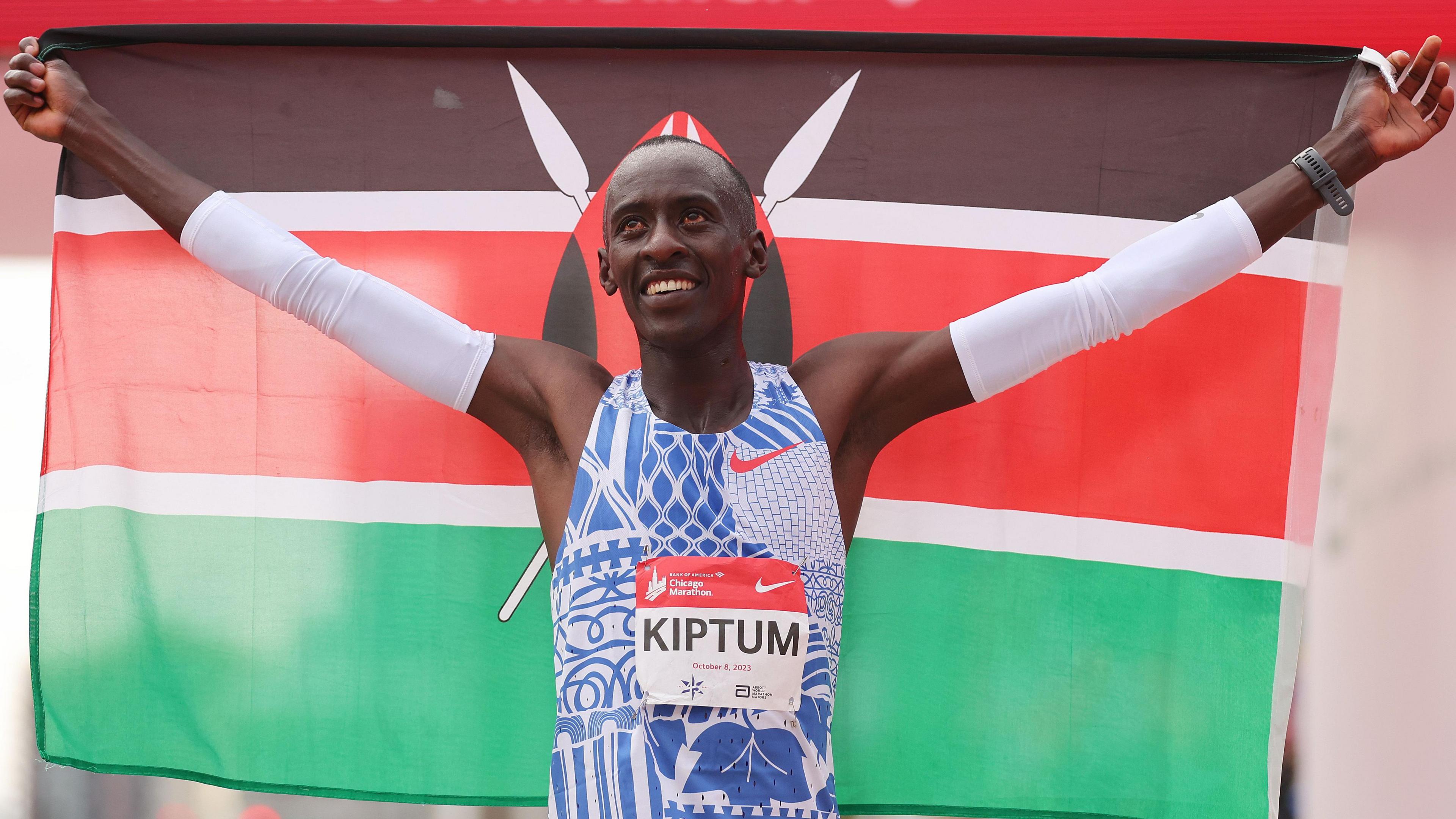 Kelvin Kiptum, wearing a blue and white running vest with his name on the front and white arm-warmers holds a Kenyan flag aloft behind him