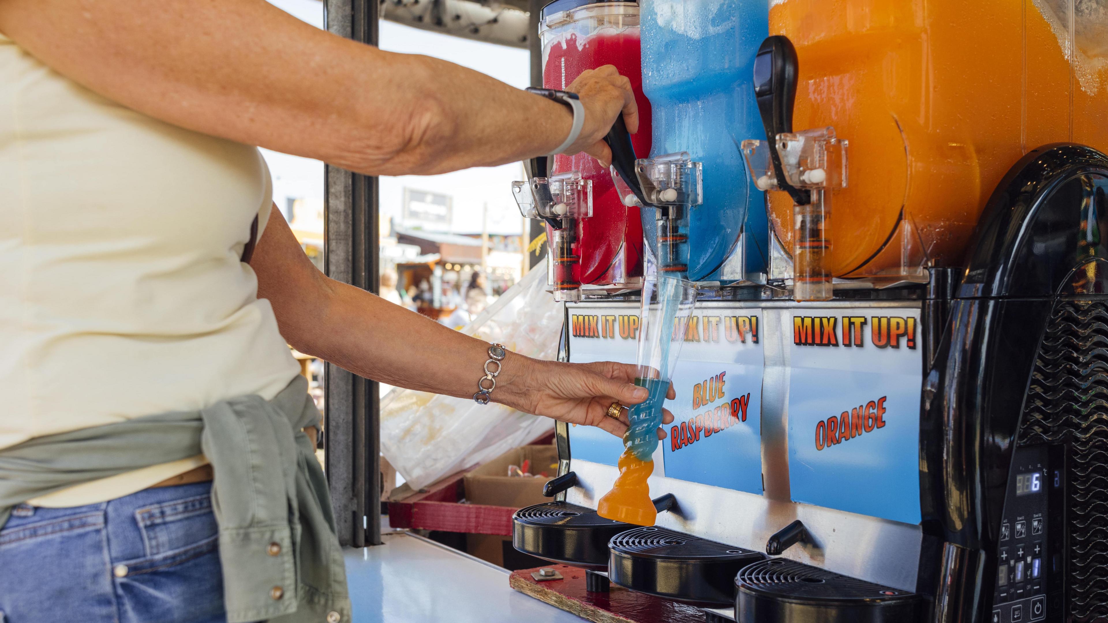 Close up off hands filling a blue slushy in a container alongside a red and orange slushy container 