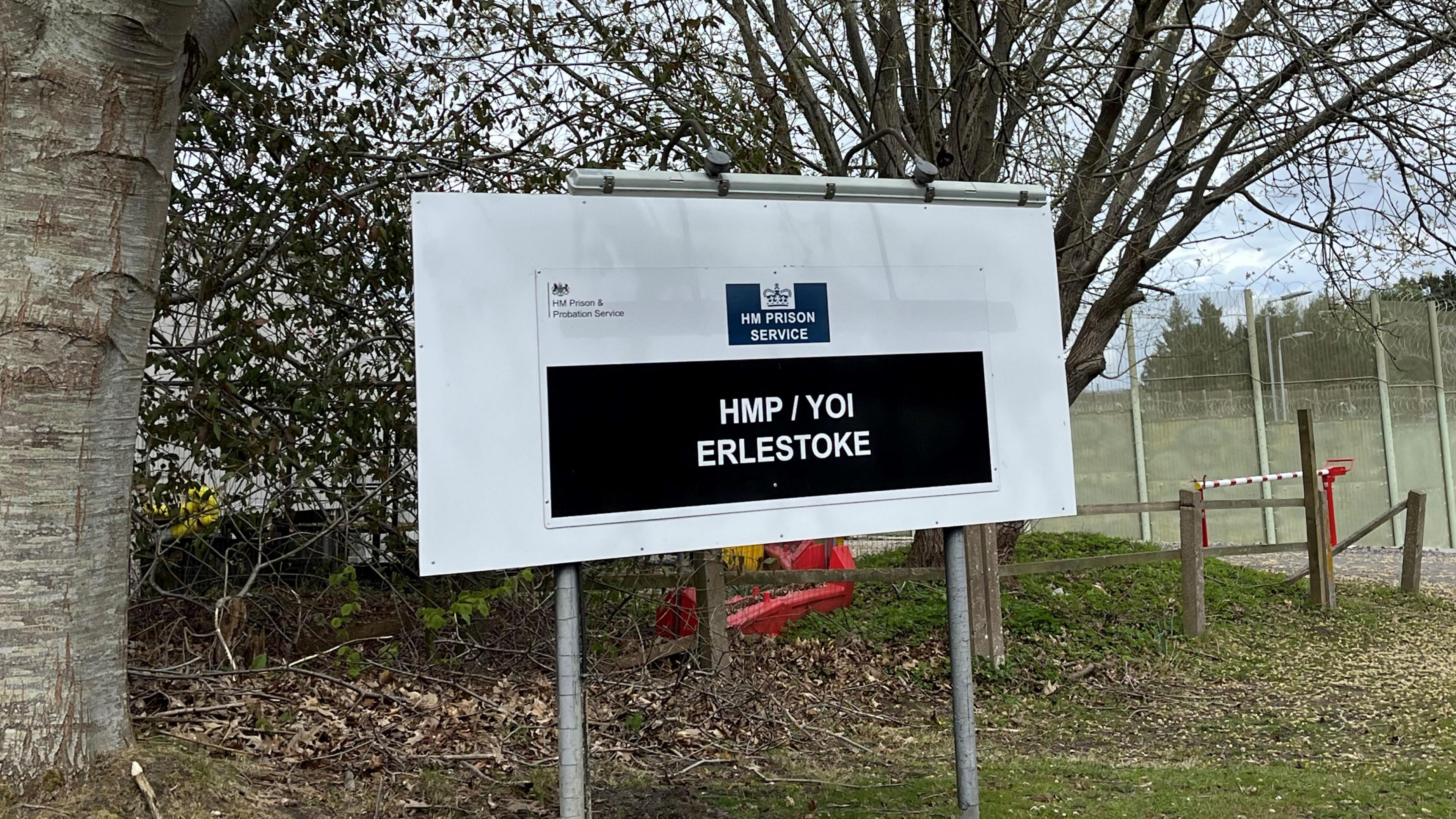 A sign outside a prison complex which reads "HM Prison Service: HMP/YOI Erlestoke". There are trees and grass behind it.