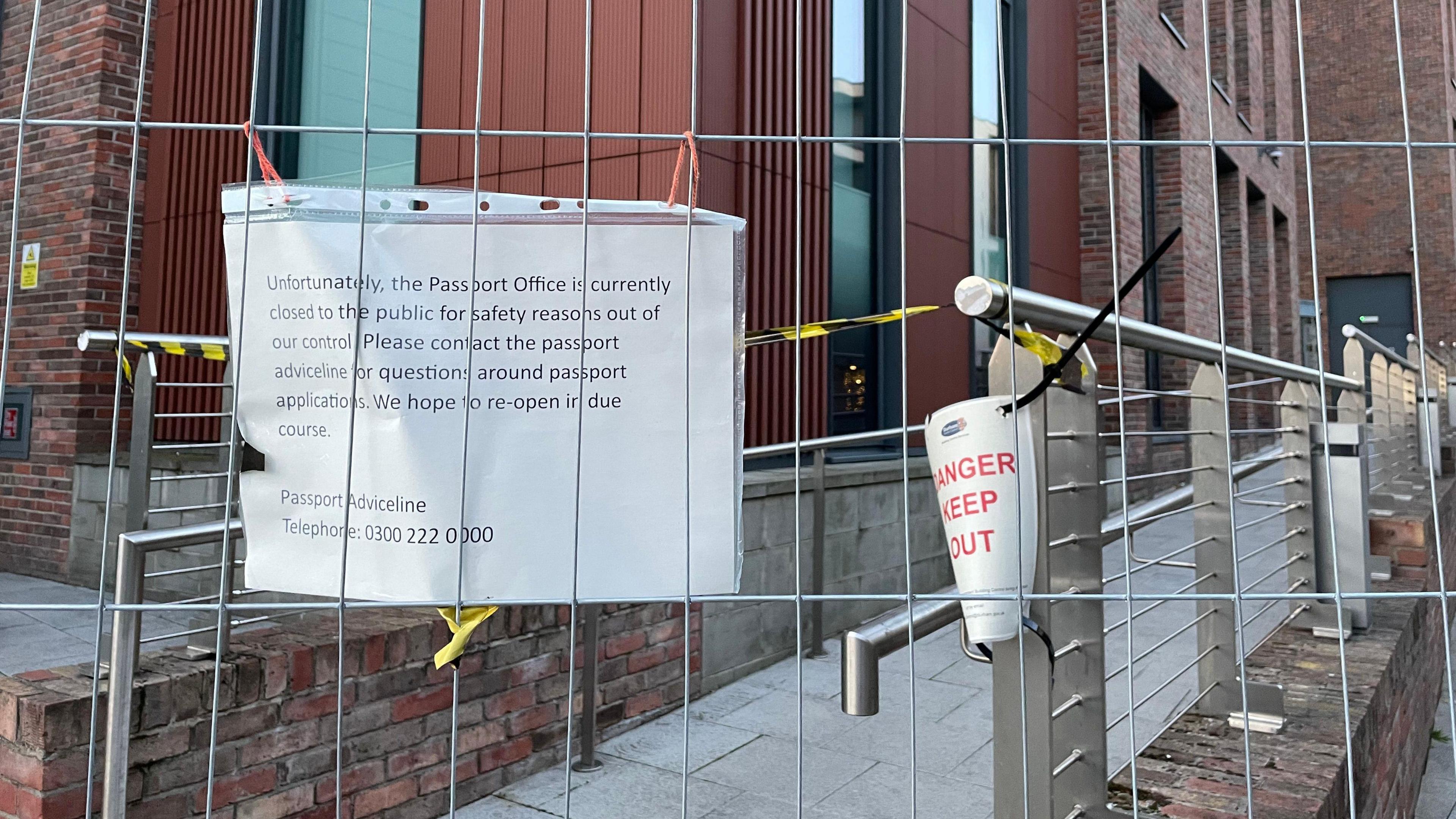 A sign attached to the metal fence which reads: "Unfortunately, the Passport Office is currently closed to the public for safety reasons out of our control. Please contact the passport adviceline for questions around passport applications. We hope to reopen in due course. Passport Adviceline Telephone: 0300 222 0000."