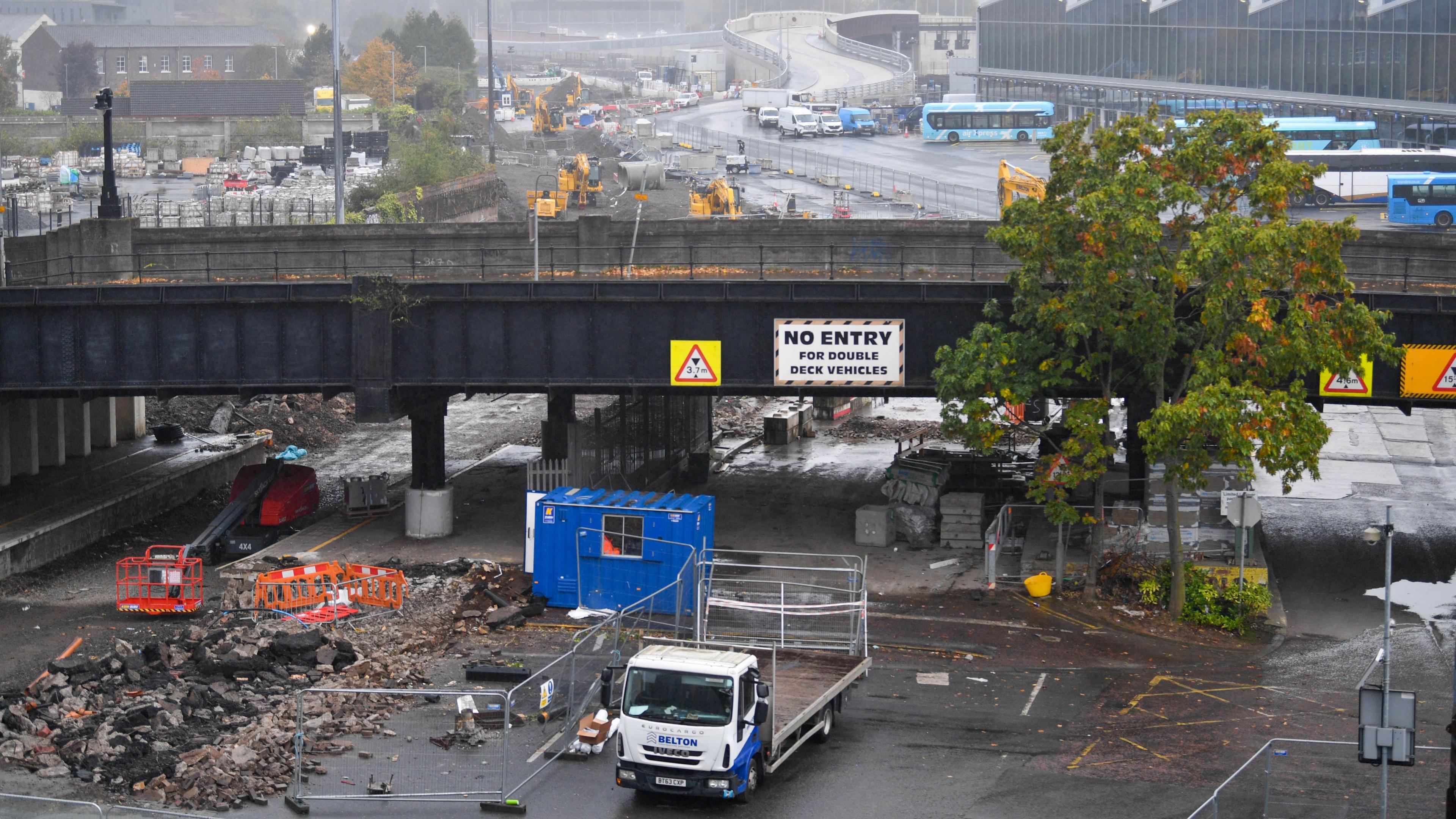 Dismantling of Boyne Bridge. There is a work van parked next to fencing. Bricks have been dug up from the ground. Grand Central Station can be seen in the background. The ground is wet.