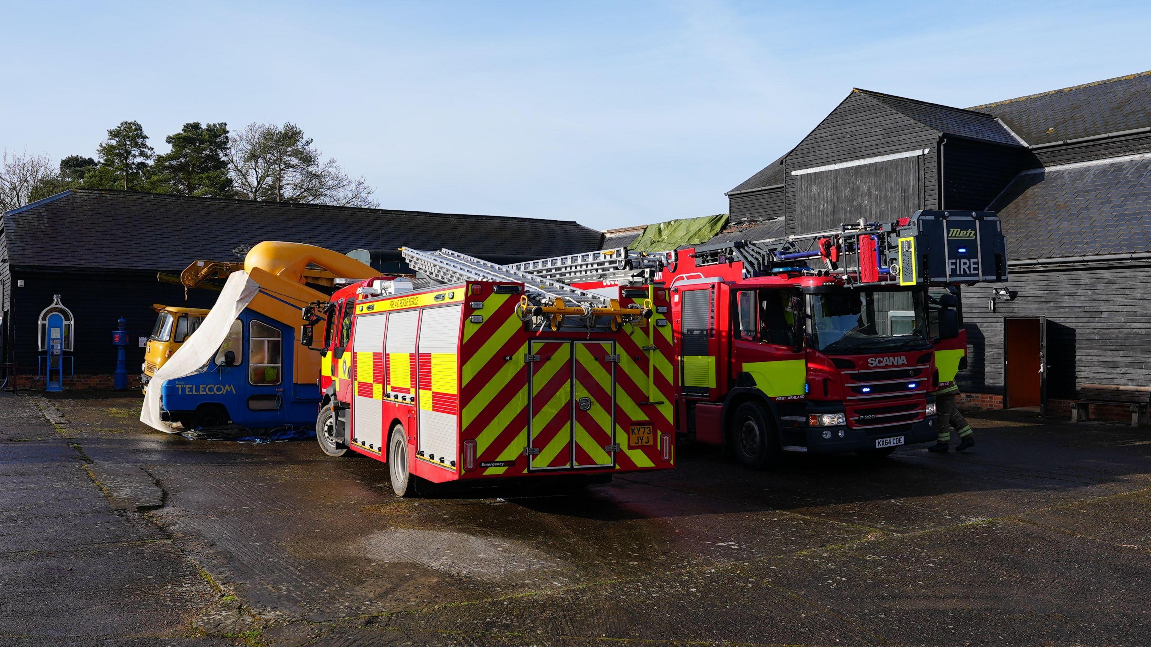 Fire engines parked behind the black timber museum building. They are parked next to an old telecom van which is blue with a large yellow phone decoration on the roof.