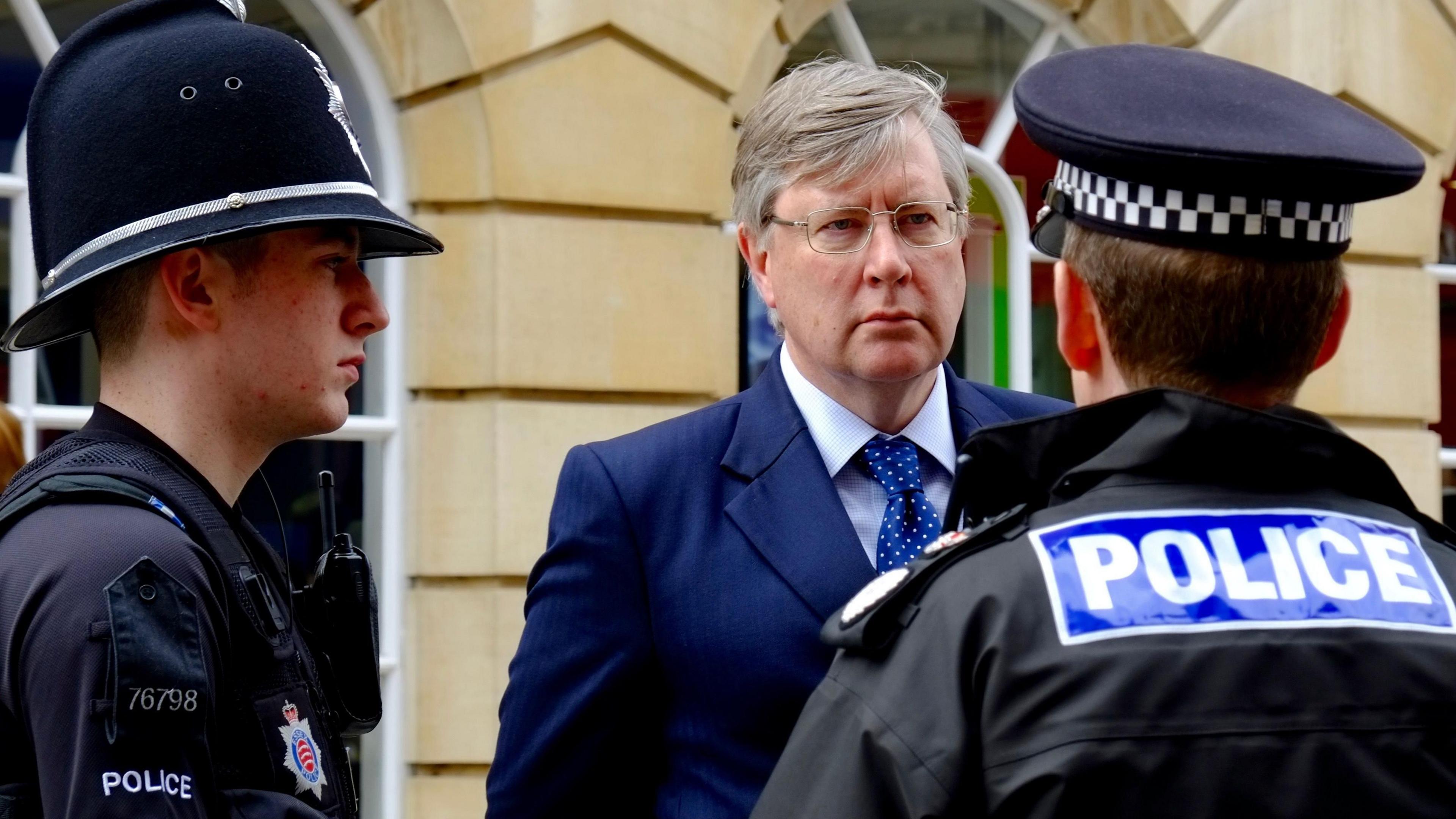 Roger Hirst is wearing a navy suit and blue tie while talking with two male uniformed police officers. He has grey hair and is wearing glasses.