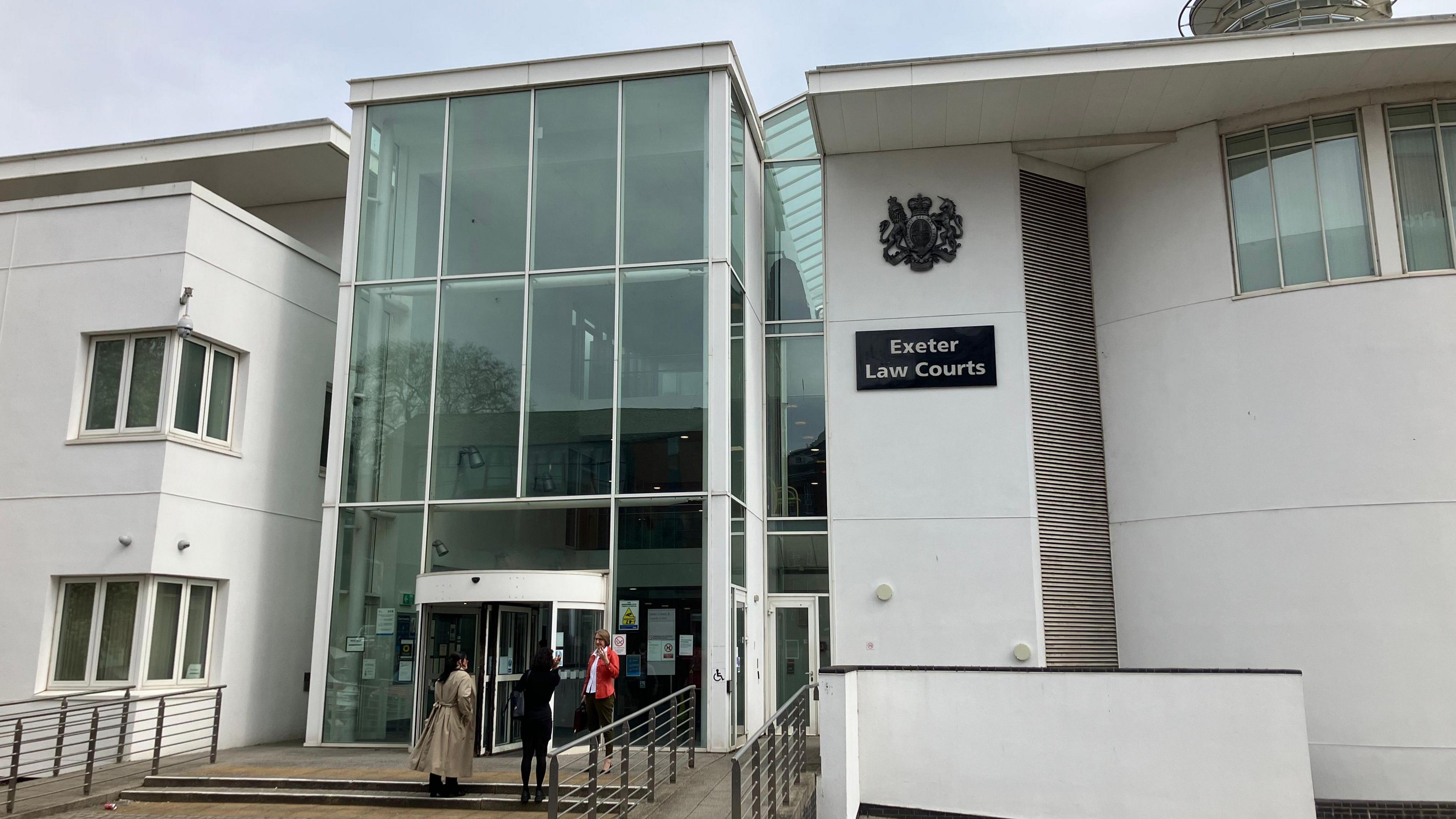 A white crown court building with a large glass entrance. A sign reads Exeter Law Courts. People stand on the steps.