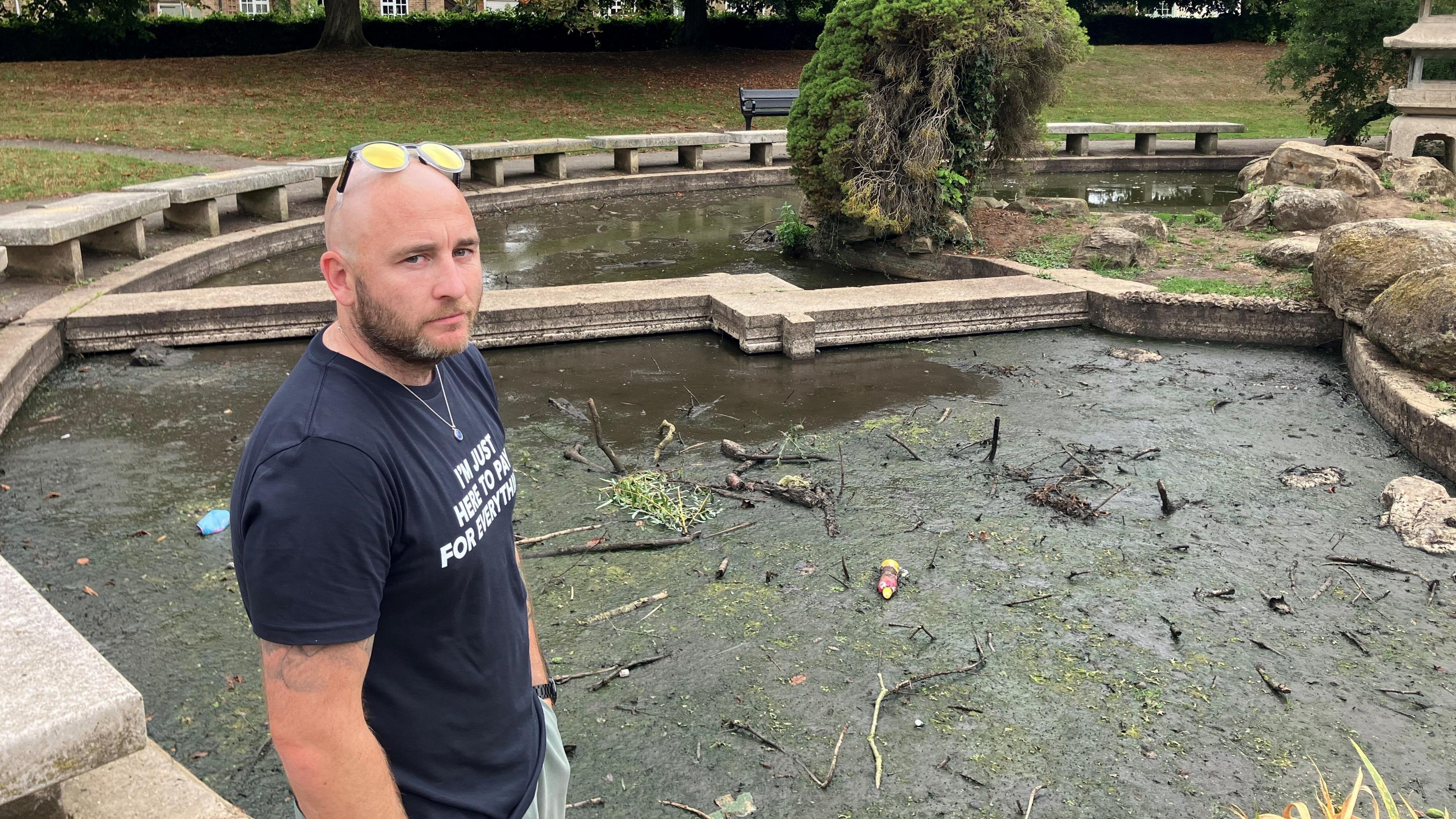 A man in a black T-shirt stands in front of a green-coloured pond with scum and branches floating on the surface.