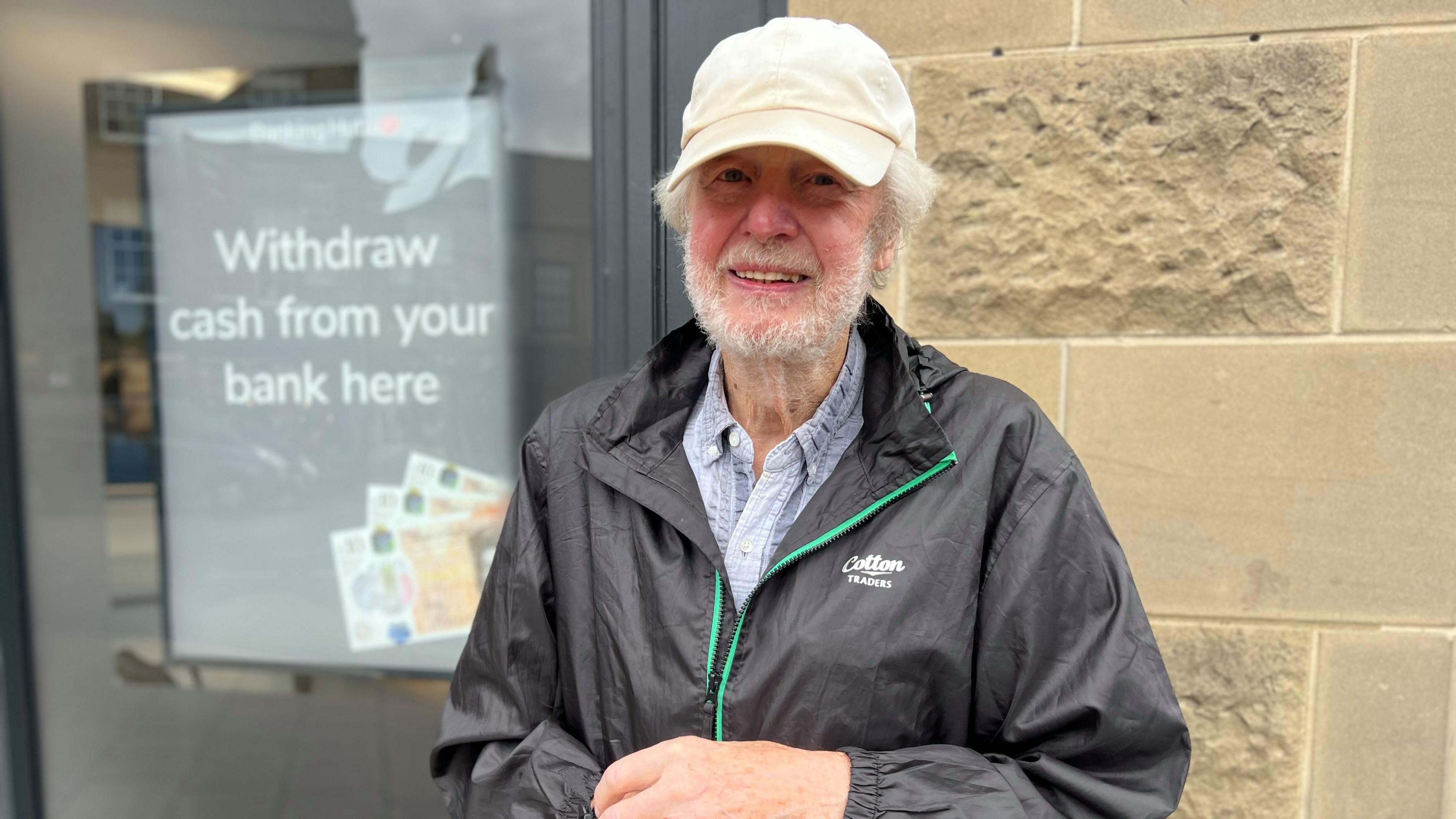 Man in cream cap with grey beard and black rain coat stood infront of brown stone building