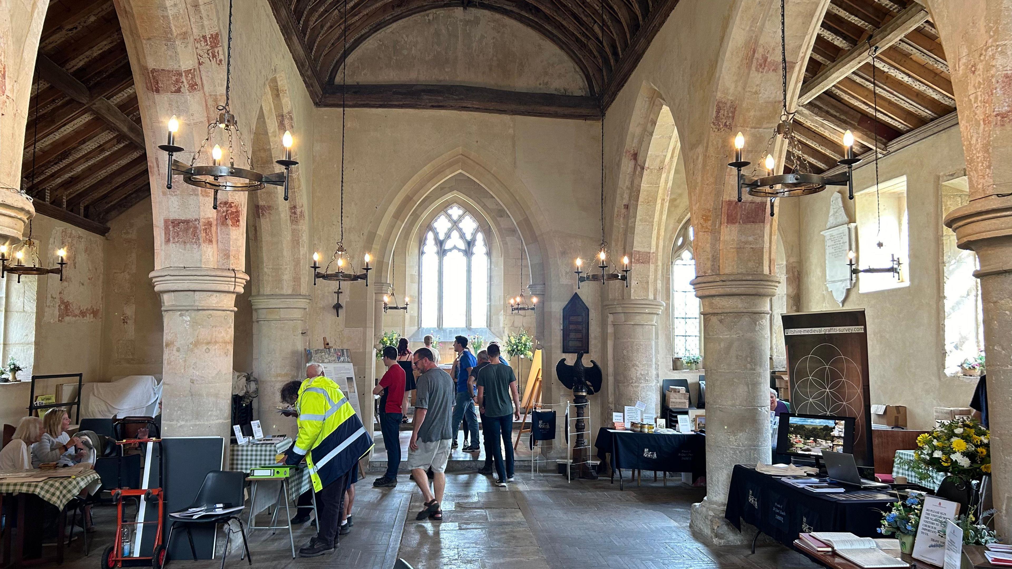 The inside of an old church - archways and pillars down both sides with tables and chairs and information stalls. The public wanders about the church,