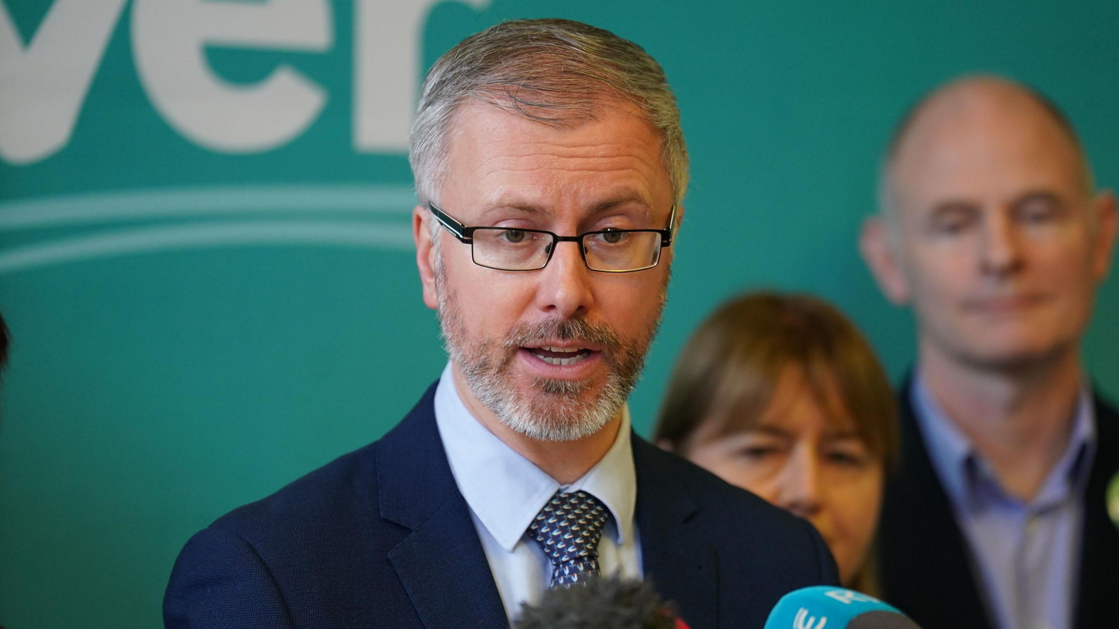 Roderic O'Gorman in a blue shirt and navy suit jacket against a green wall, speaking into microphones with party members in the background