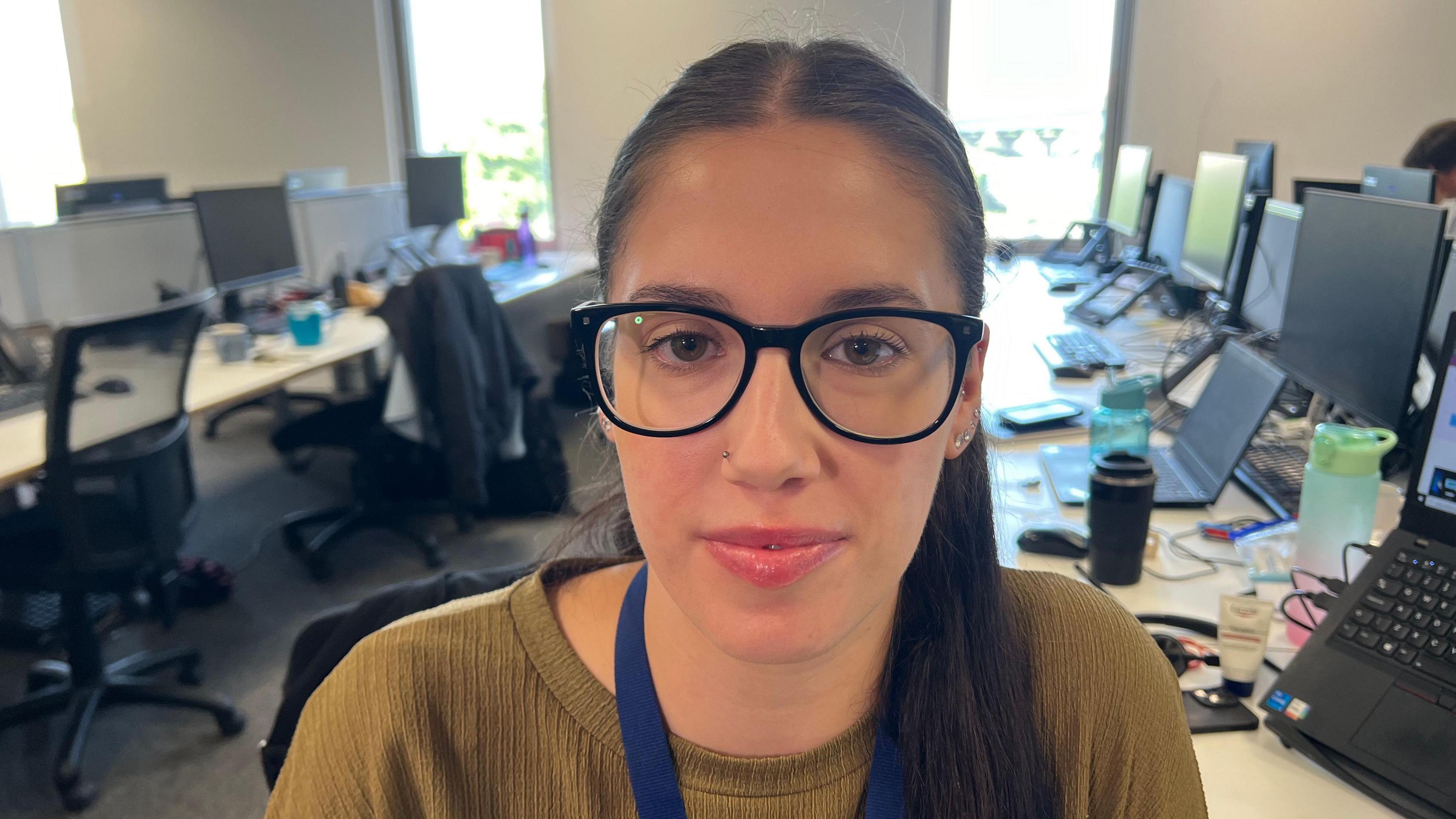 A woman wearing a olive-green top and glasses in an office, looking at the camera