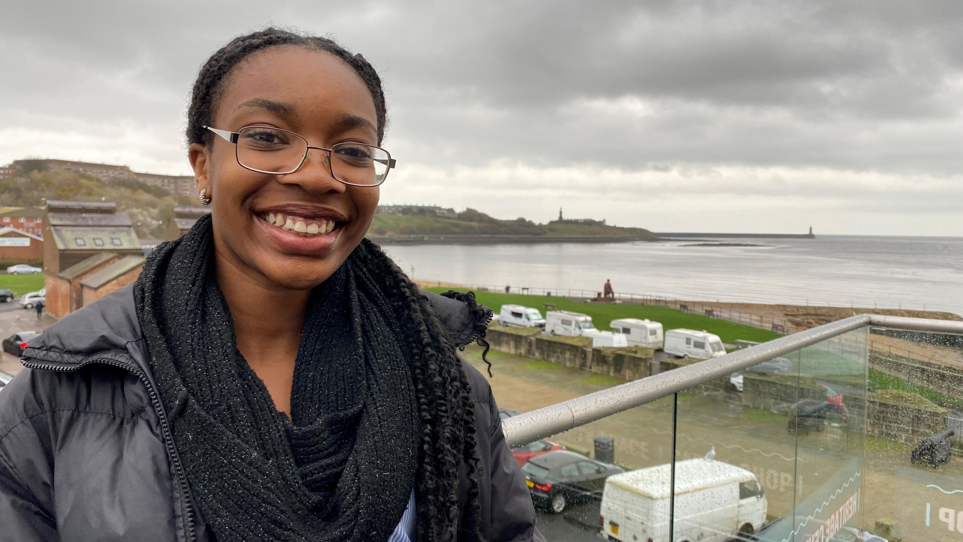 Deborah is standing on a balcony with the River Tyne behind her. She's a teenager with her hair in braids and wearing a black coat and scarf.   