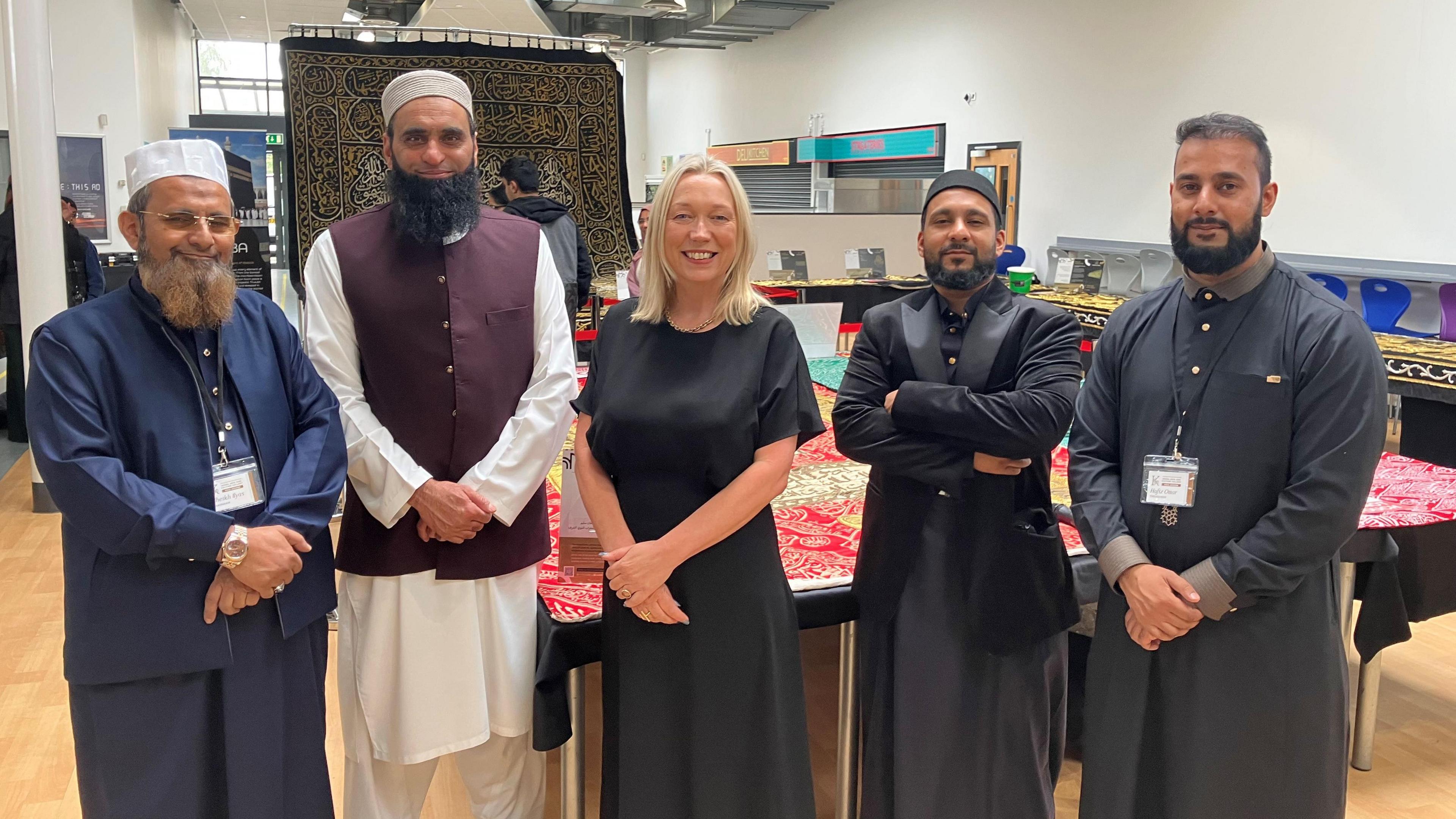 Four men in Islamic clothing and with beards with one woman with shoulder length blonde hair and a dark dress. Behind them are rare Islamic artefacts in a school hall.