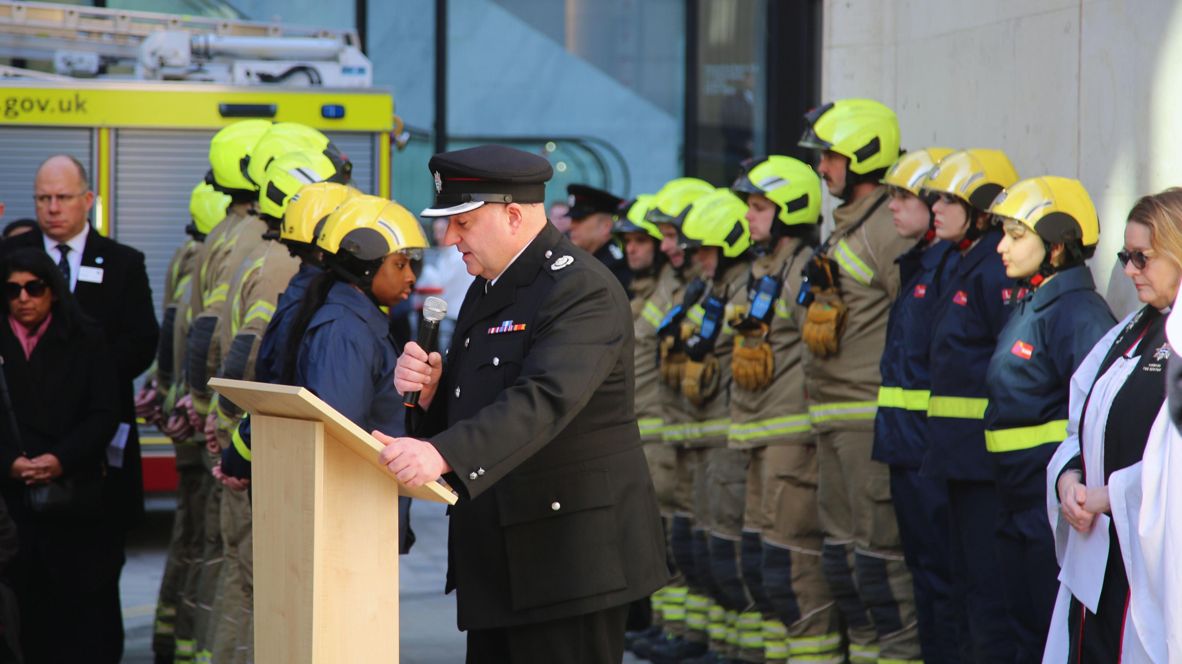 A man wearing a black uniform with a cap on holding a microphone and reading. There are members of the fire brigade stood behind him in a line. 