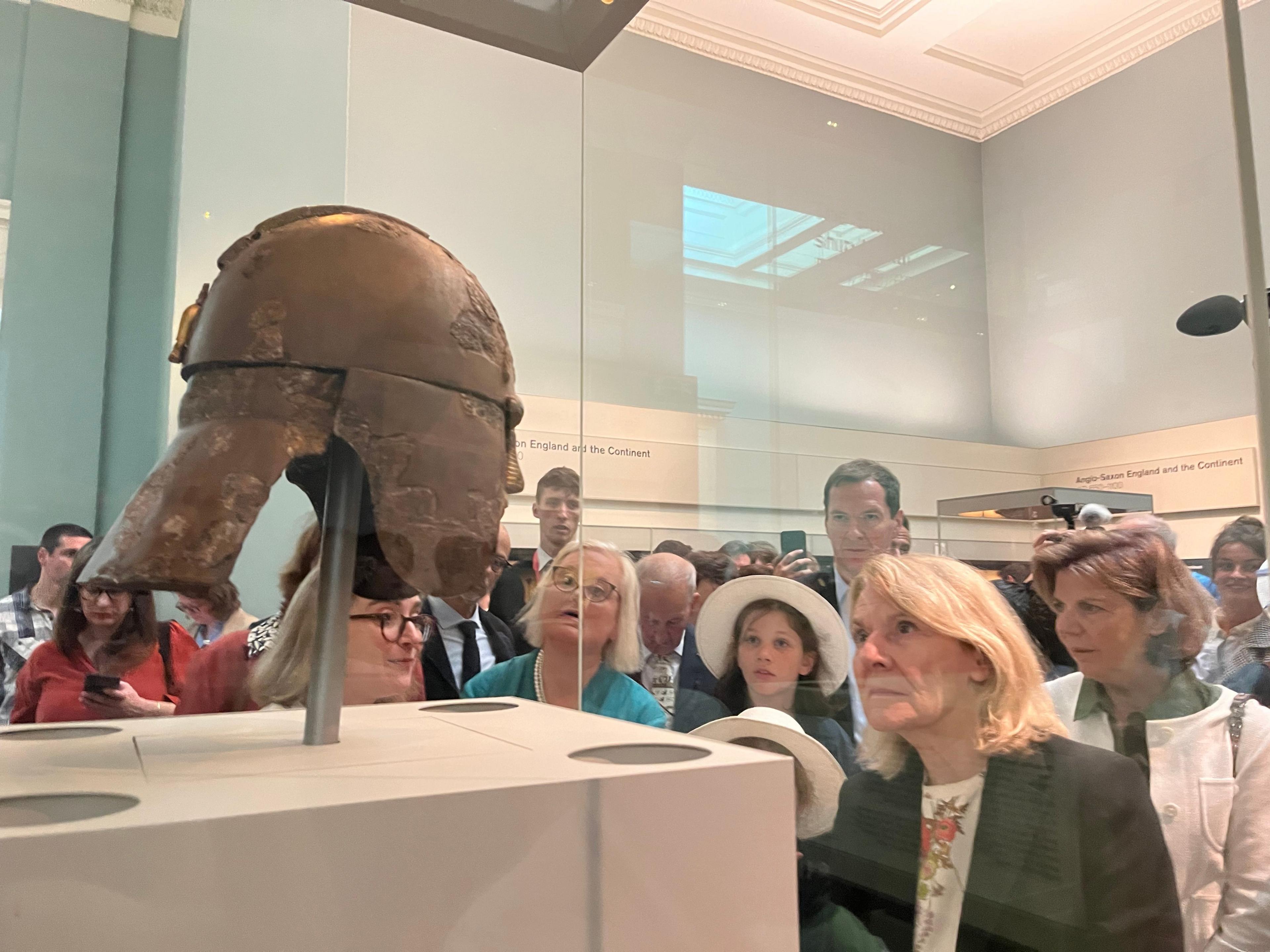 Side view of the iron Sutton Hoo Helmet in a glass case with French Culture Minister in blonde hair looking intently at it -- with Osborne behind looking on